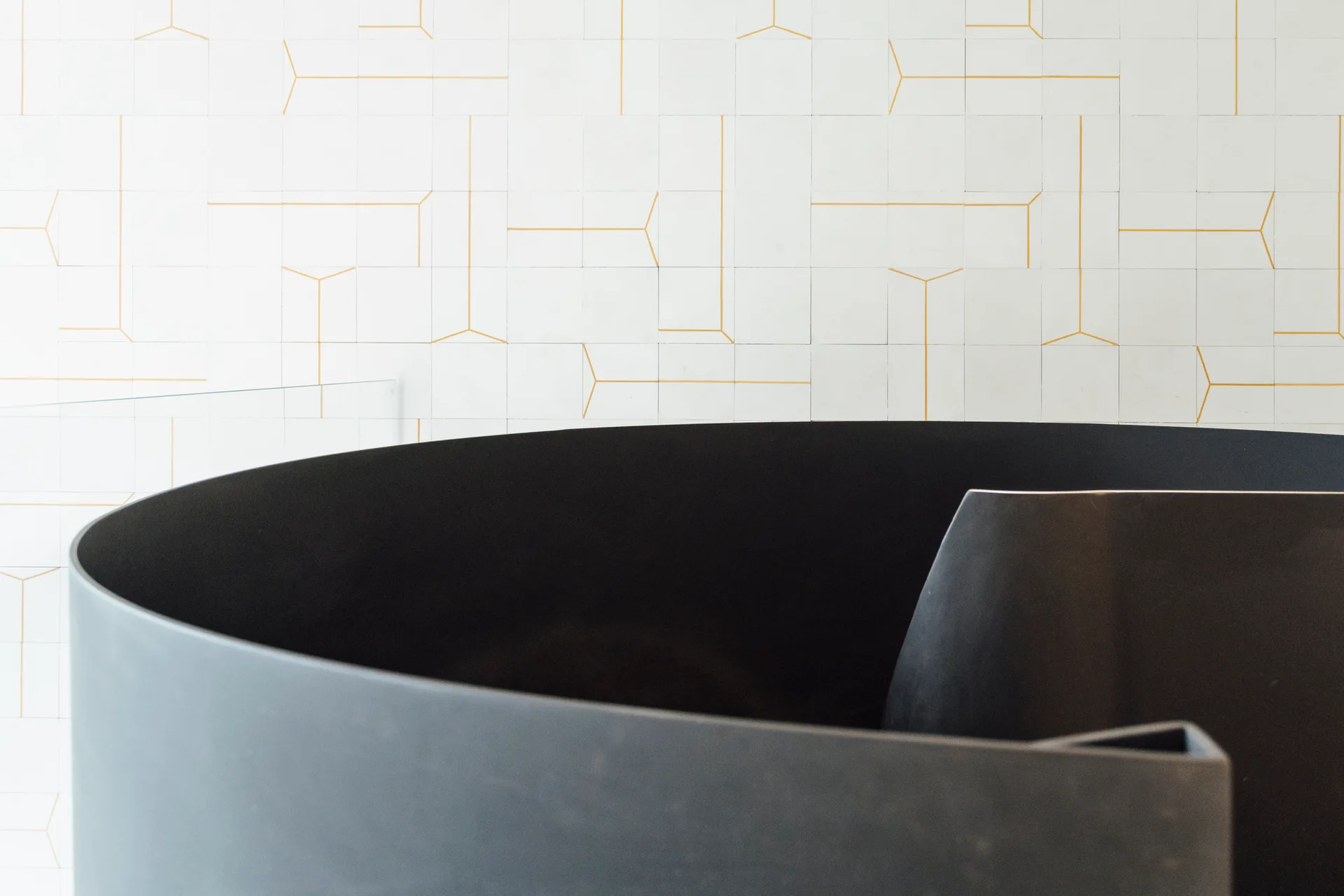 A close-up of a matte black curved stair railing positioned against a white tiled wall featuring thin gold geometric lines.