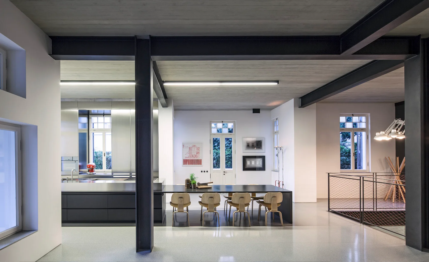 An industrial kitchen and dining space with exposed black steel columns, a concrete ceiling, a long dark gray island, and wooden chairs.