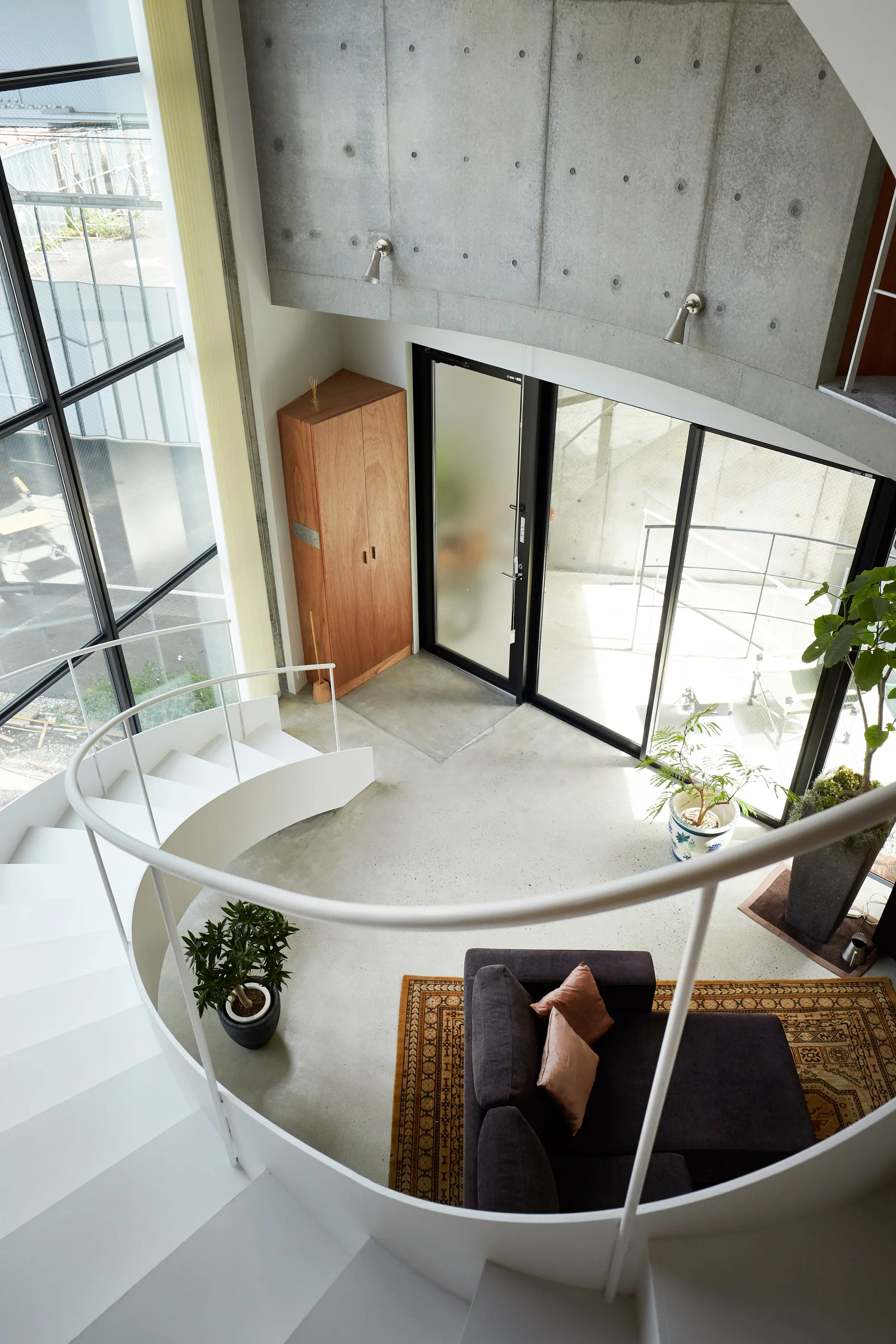 A high-angle view down a curved white staircase into a minimalist living area with exposed concrete walls, a dark sofa, and a patterned rug.