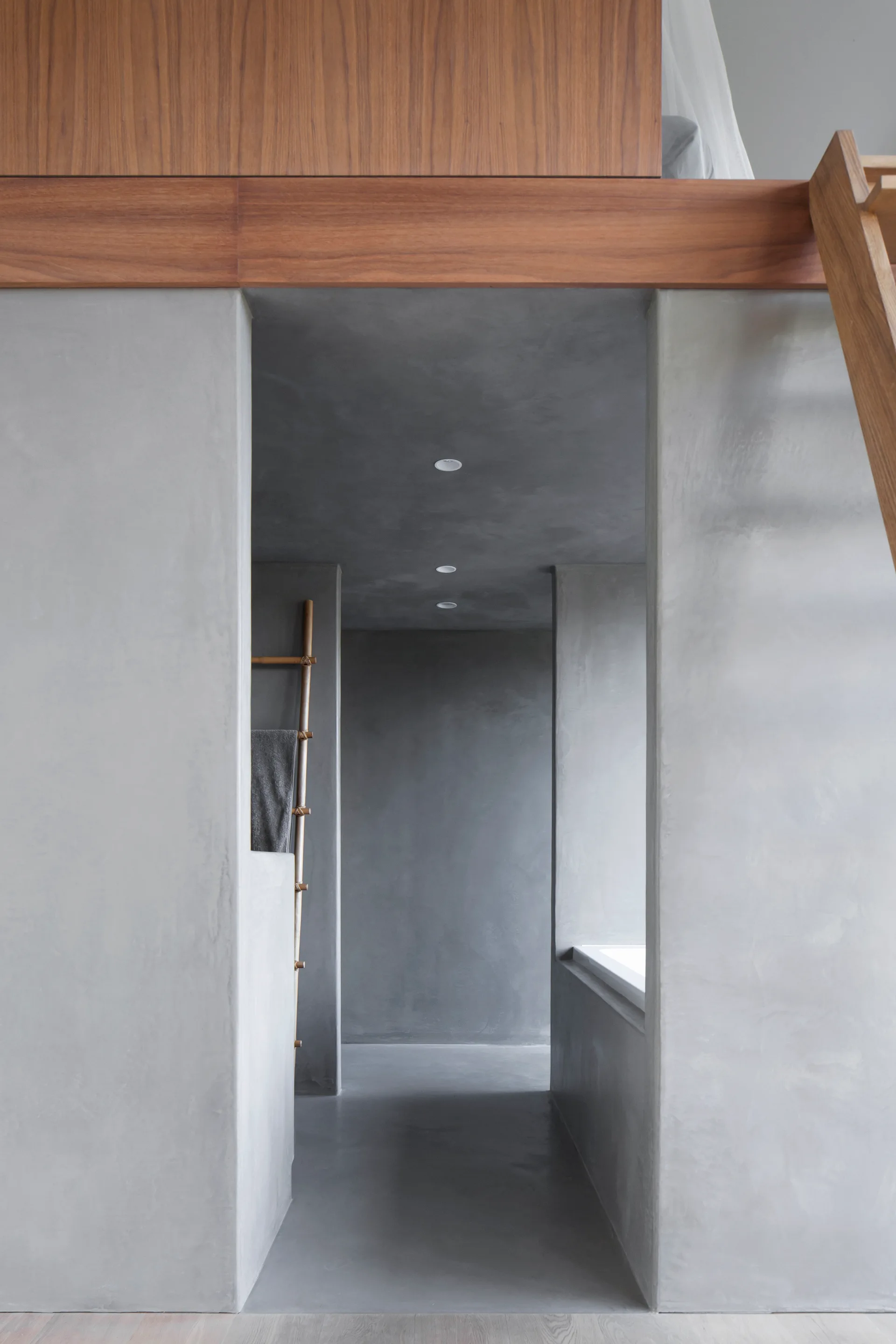 A minimalist bathroom interior features seamless gray plaster walls and floors with a wooden ceiling beam, recessed lighting, and a towel ladder.