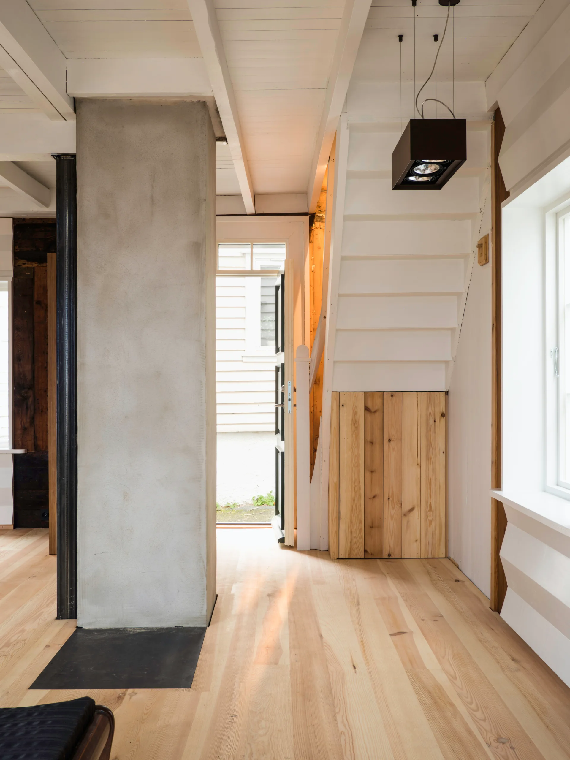 A light-filled entryway features a white wooden staircase, light-colored wood flooring, and a large raw concrete pillar.