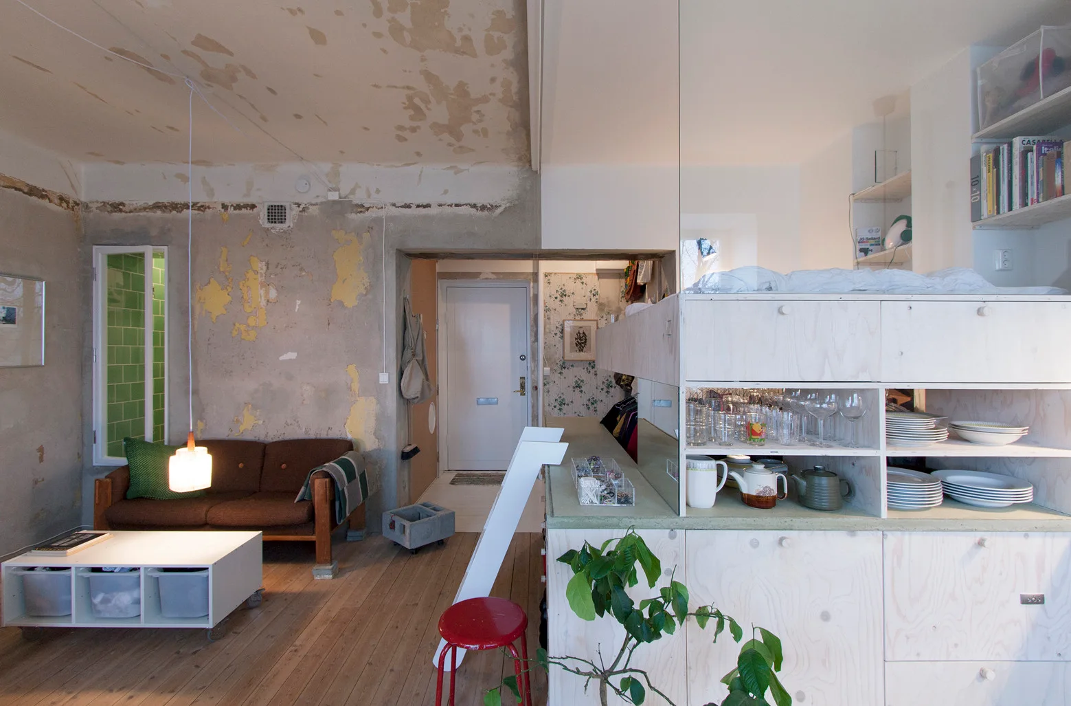 An industrial living space features distressed plaster walls, a brown sofa, a low white coffee table, and a large plywood room divider with open shelves.