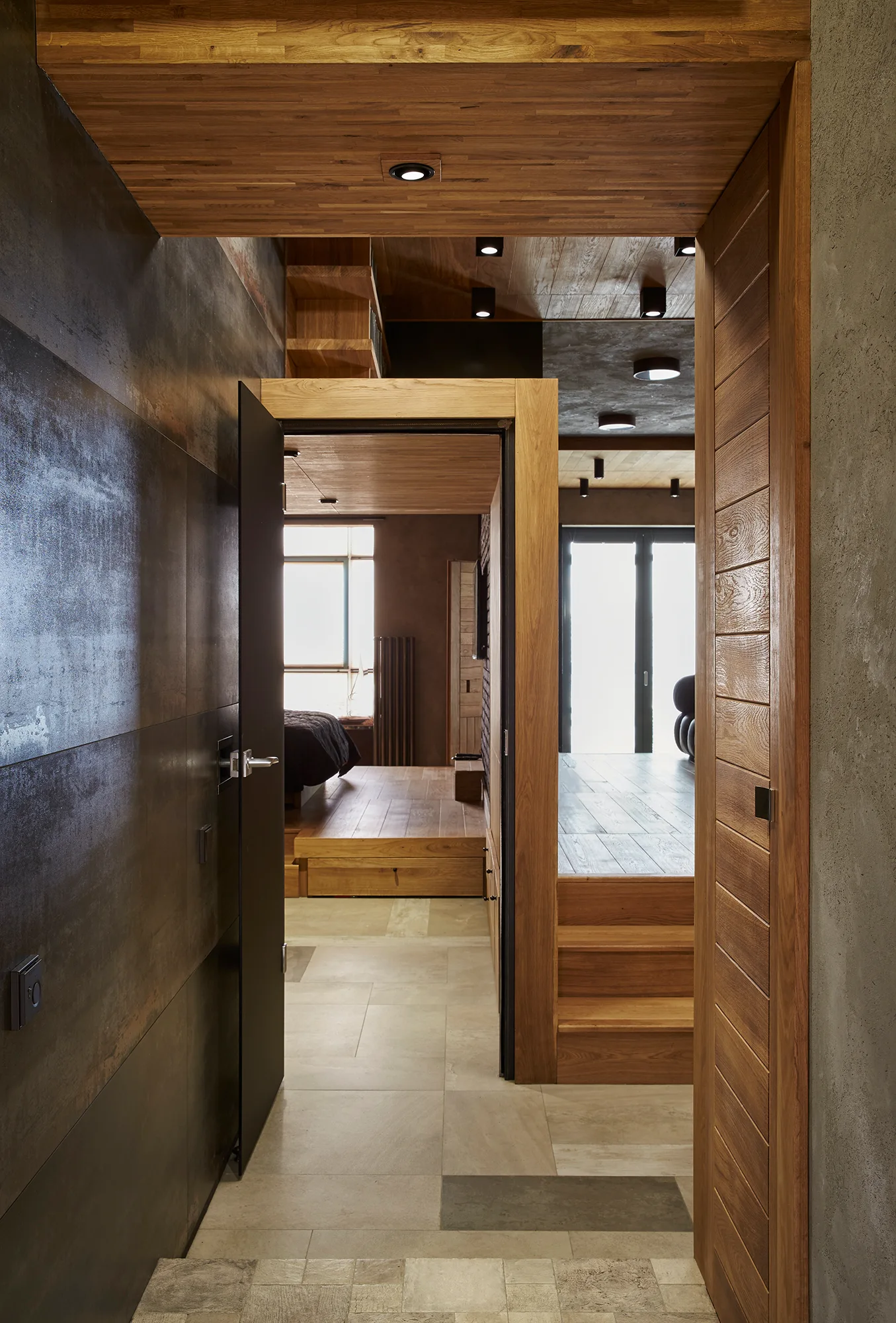A modern hallway features dark metal-look wall panels, a wood-paneled ceiling, and steps leading to elevated wooden platform rooms.