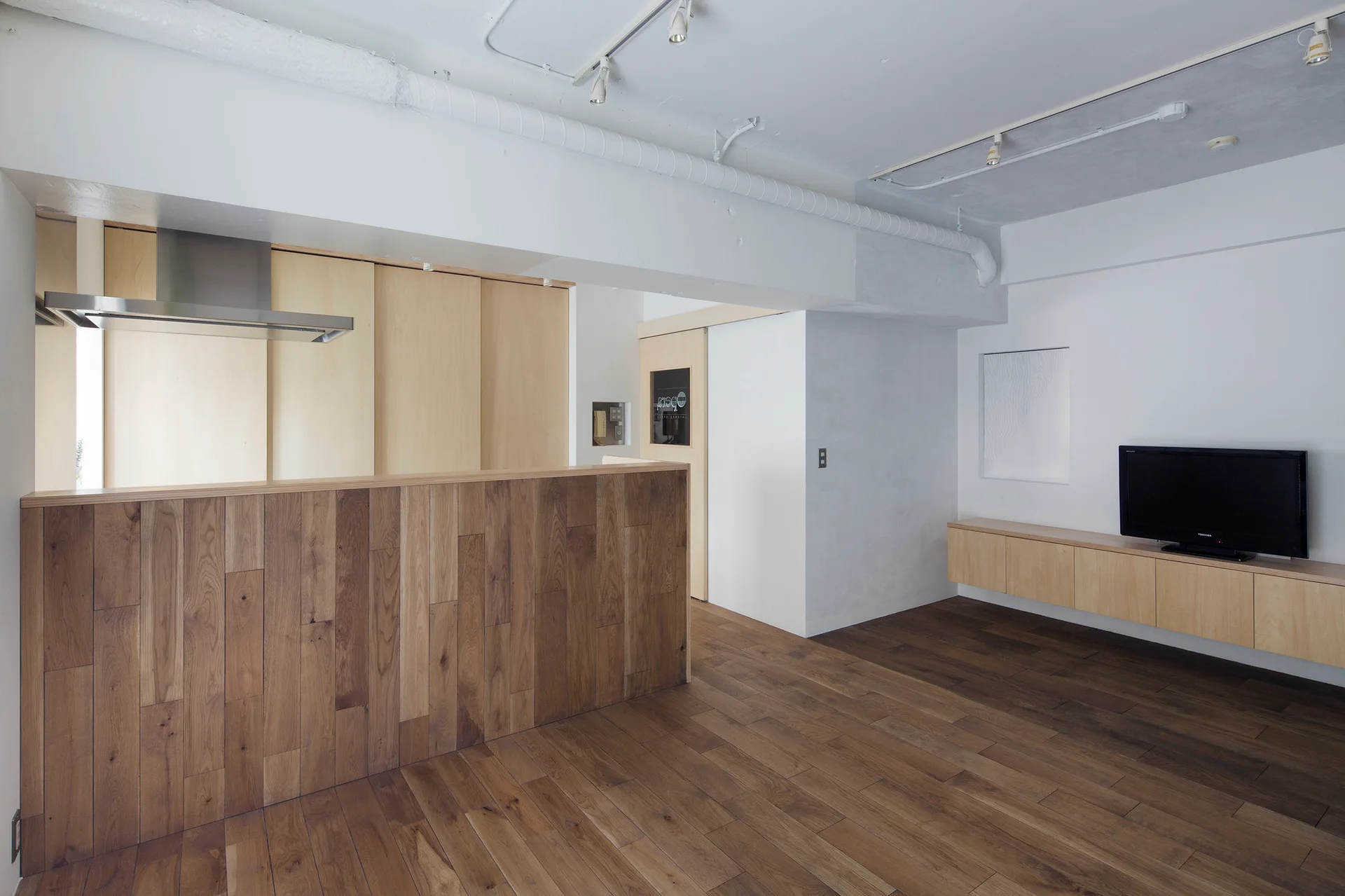 A minimalist open-plan living room features a wood-paneled kitchen island, matching wood floors, a floating media console, and white track lighting.