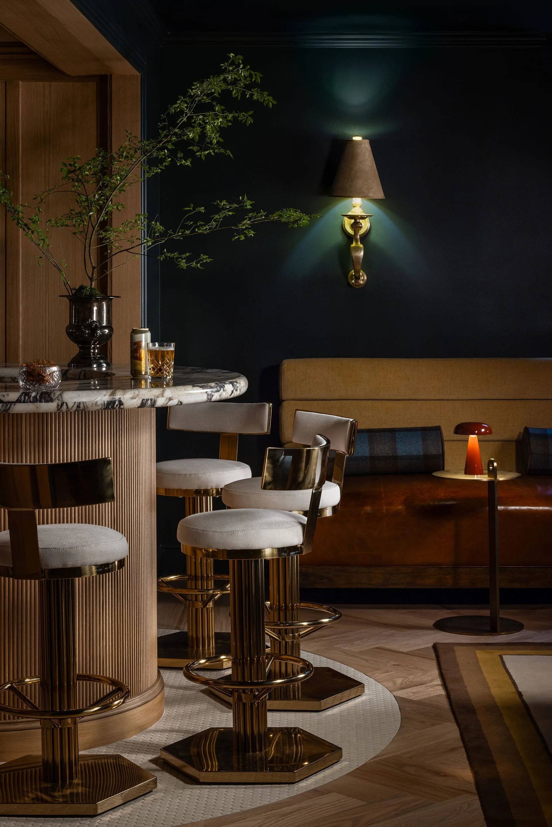 A curved marble-topped bar with a fluted wooden base and brass stools stands before a dark blue wall and a leather banquette.