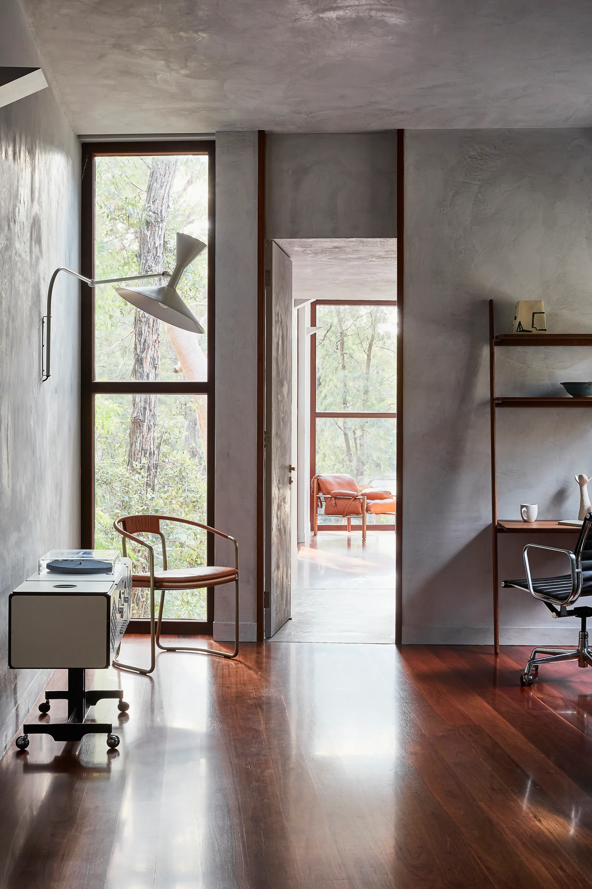 A home office featuring textured gray plaster walls, dark polished wood floors, a mid-century desk, and a tall window with forest views.