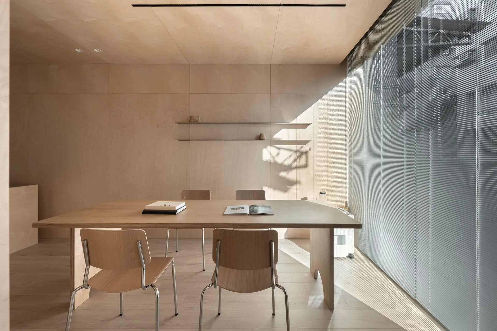 A minimalist dining room features light-toned plywood paneled walls, a matching wood table with chairs, and natural light streaming through blinds.