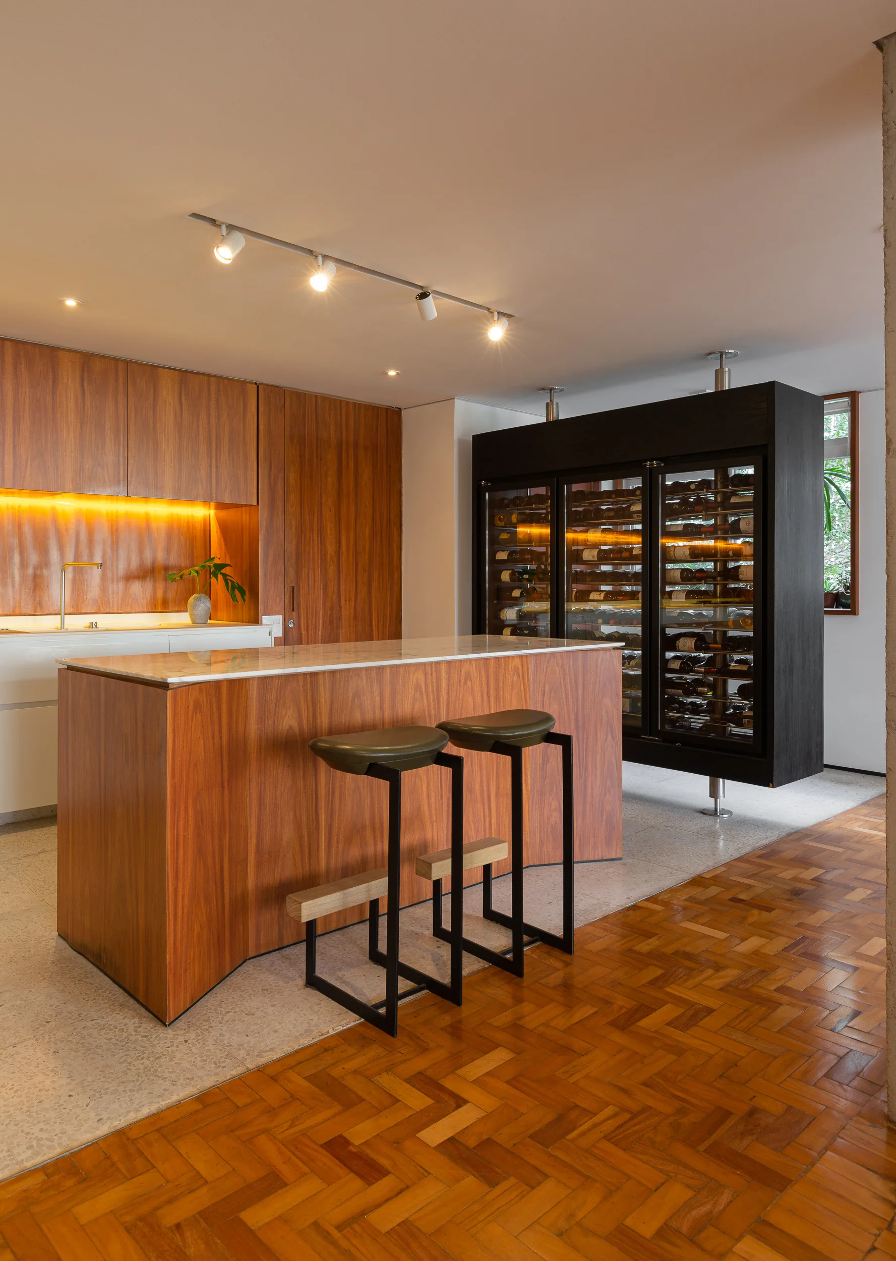 A contemporary kitchen features a wood-wrapped island with two bar stools, warm wood cabinets, and a large black wine cooler.