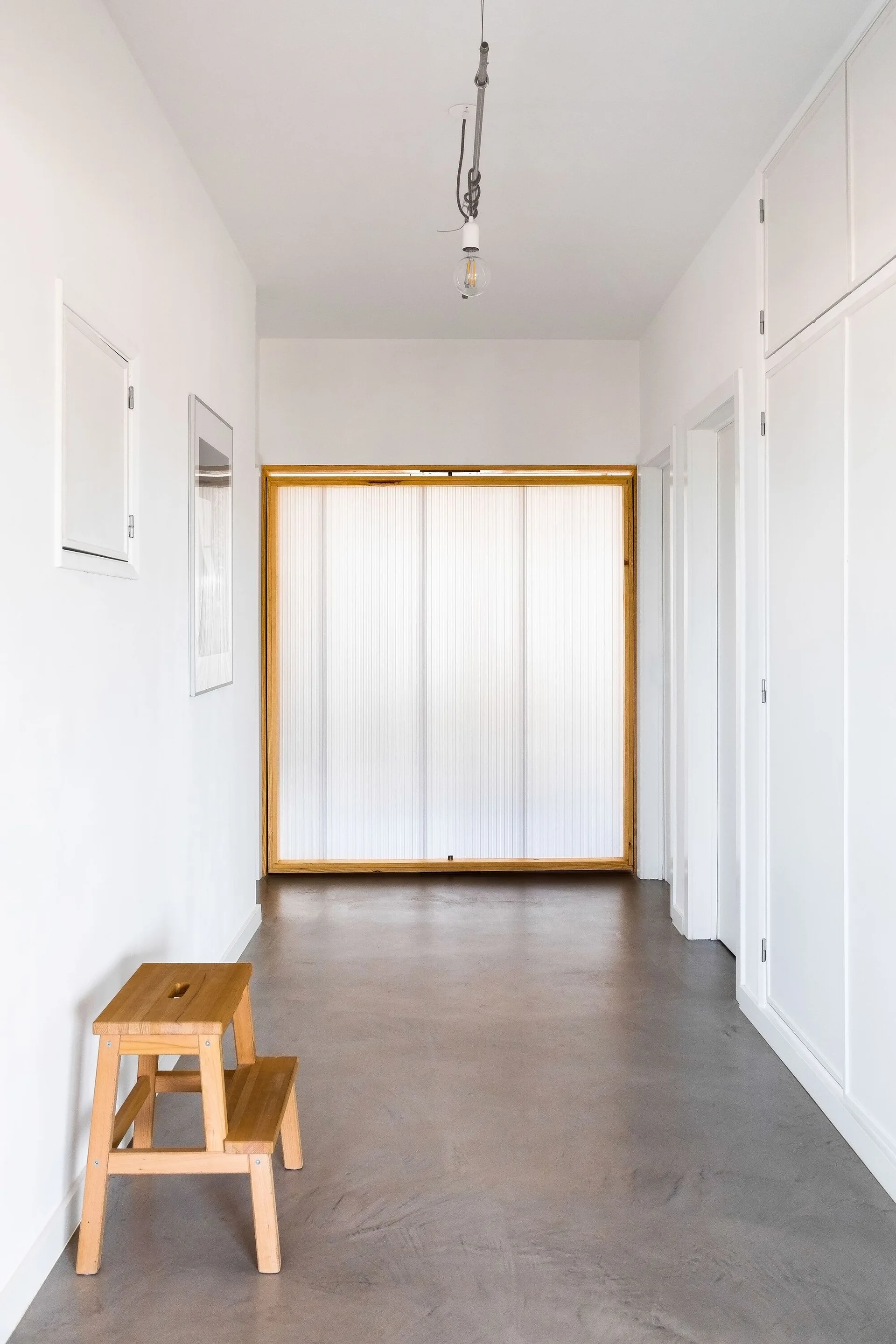 A minimalist hallway features white walls, a gray concrete floor, a wooden step stool, and a frosted glass sliding door framed in wood.