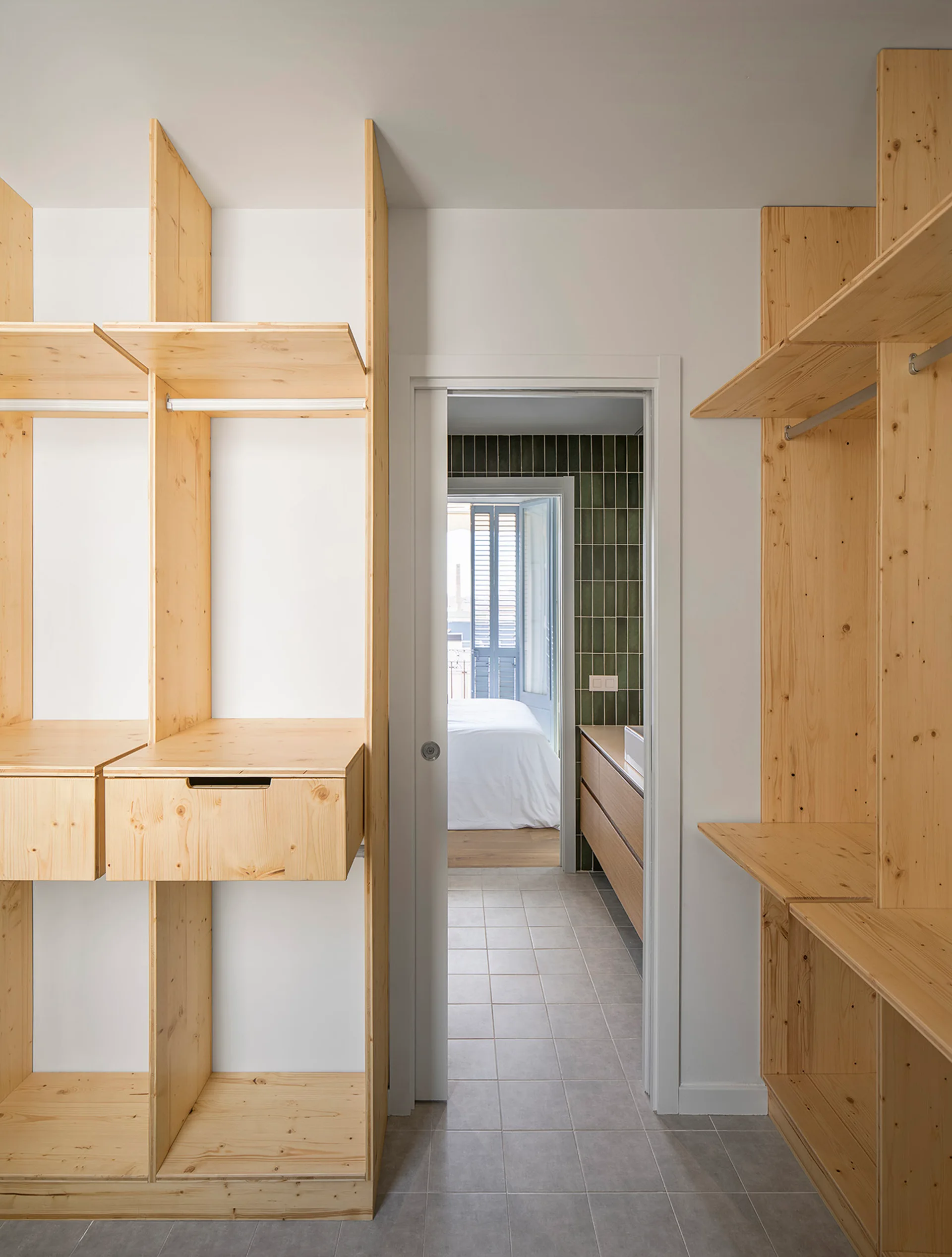 A light wood walk-in closet with built-in open shelving units leads through a sliding pocket doorway into a bathroom with grey floor tiles.