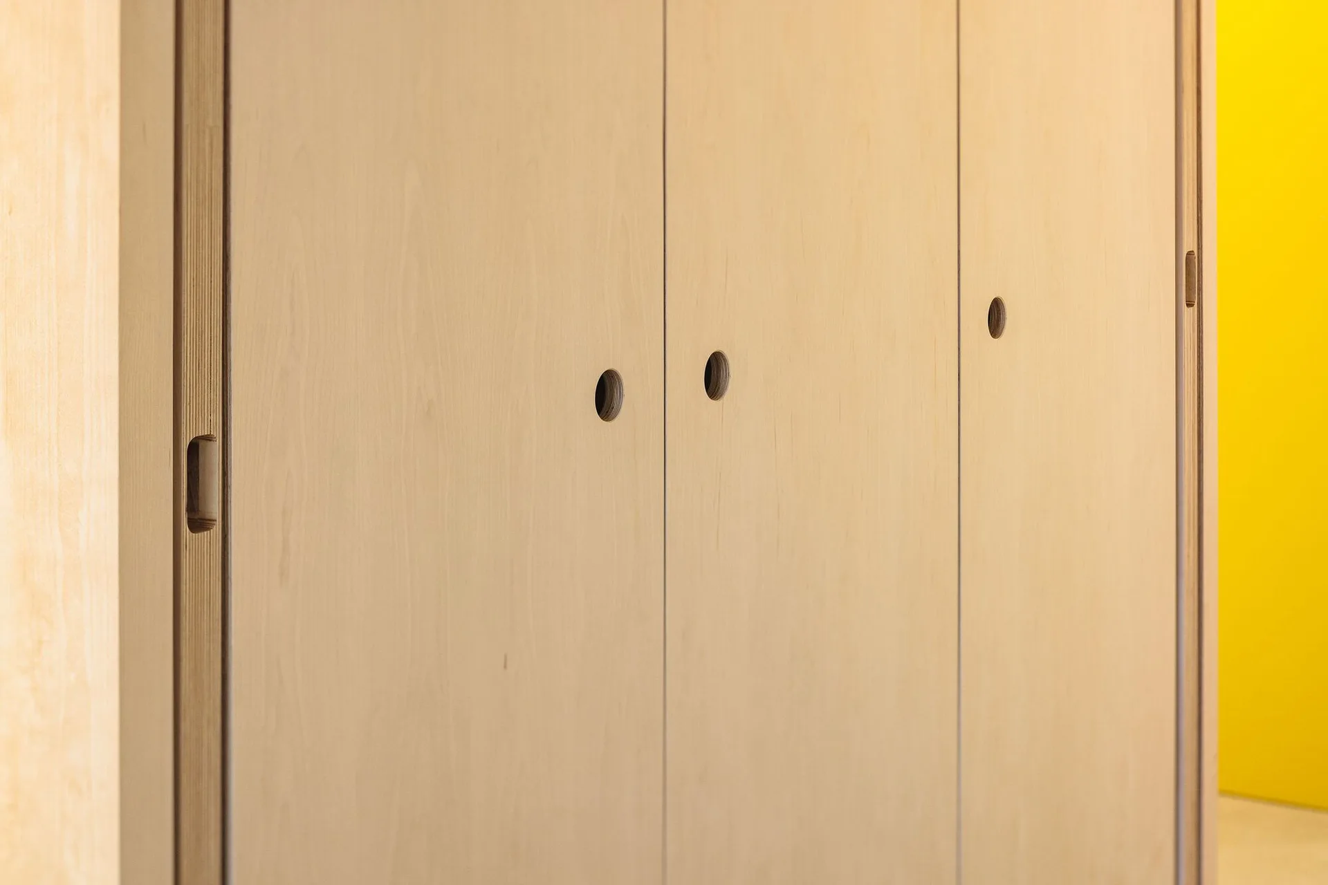 A close-up of flat-front light plywood cabinet doors featuring circular cutout finger pulls next to a bright yellow wall.