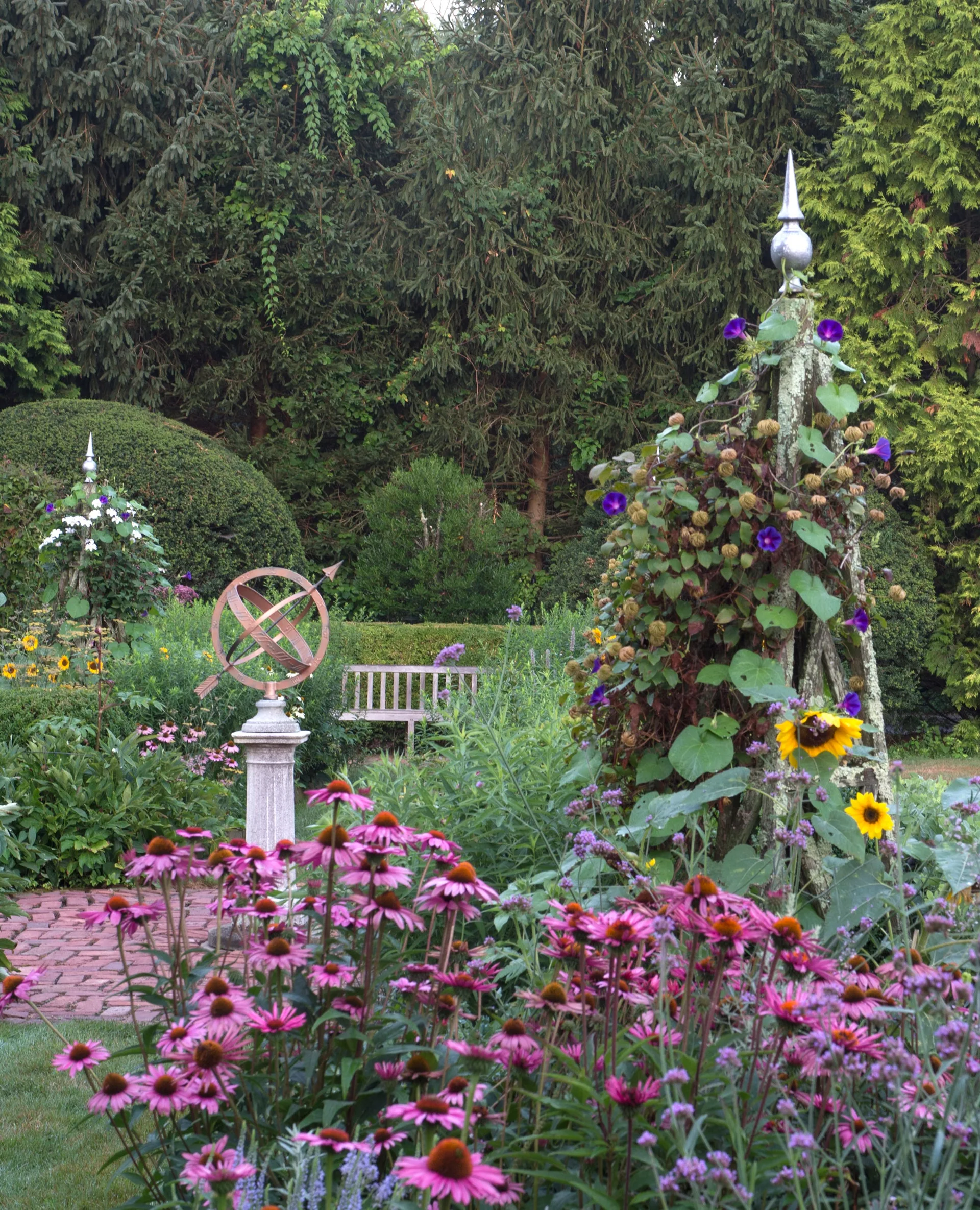 A cottage garden with purple coneflowers in the foreground, an armillary sundial on a stone pedestal, and a wooden bench near a brick path.
