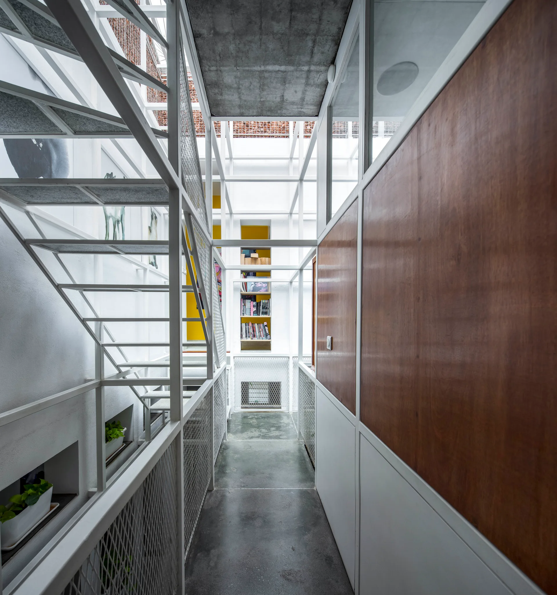 An industrial hallway features a white metal open-riser staircase with mesh railings, wood-paneled walls, a concrete floor, and a recessed yellow bookshelf.