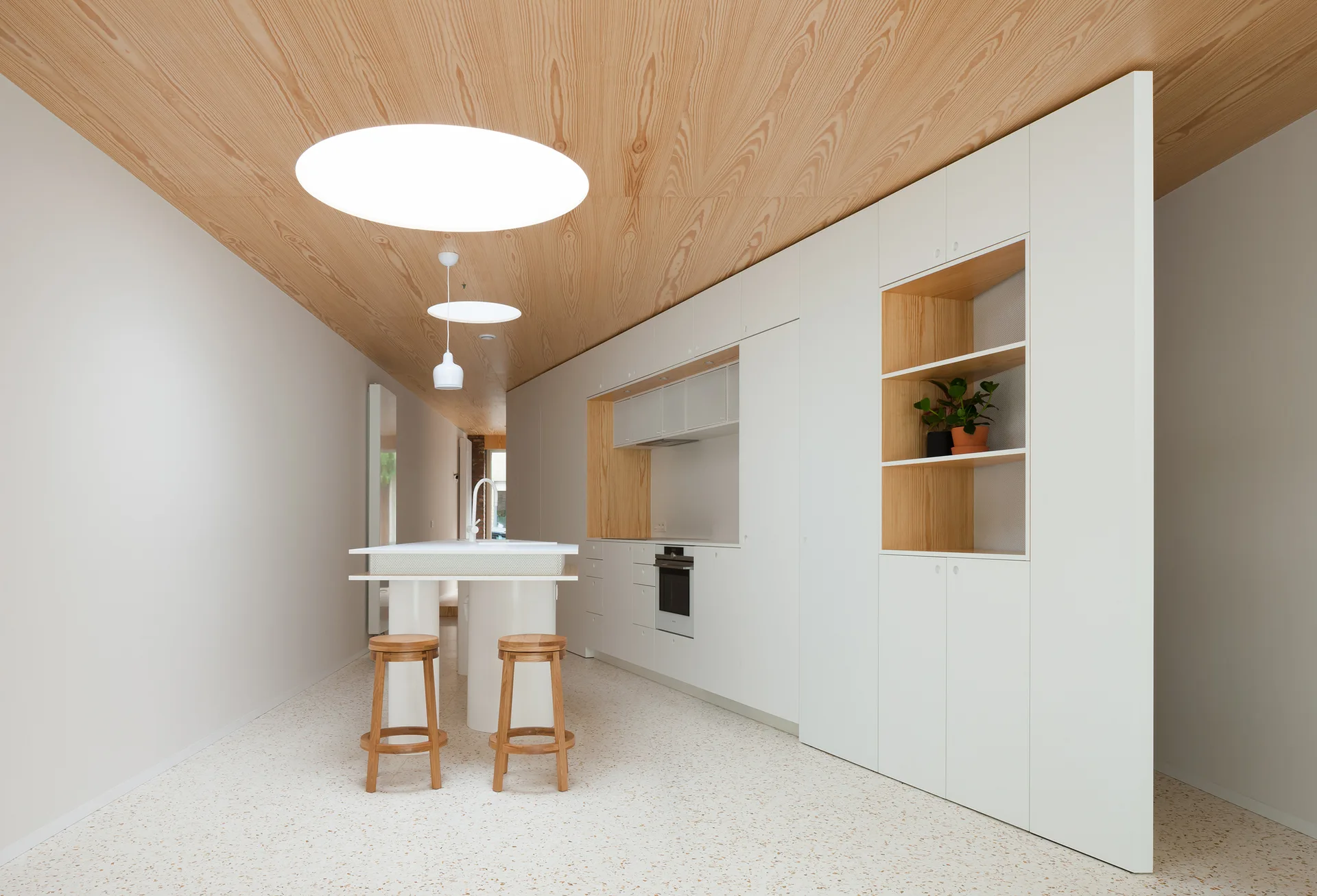 A minimalist white kitchen features a light wood-paneled ceiling with circular lights, integrated white cabinetry, a small island, and terrazzo flooring.