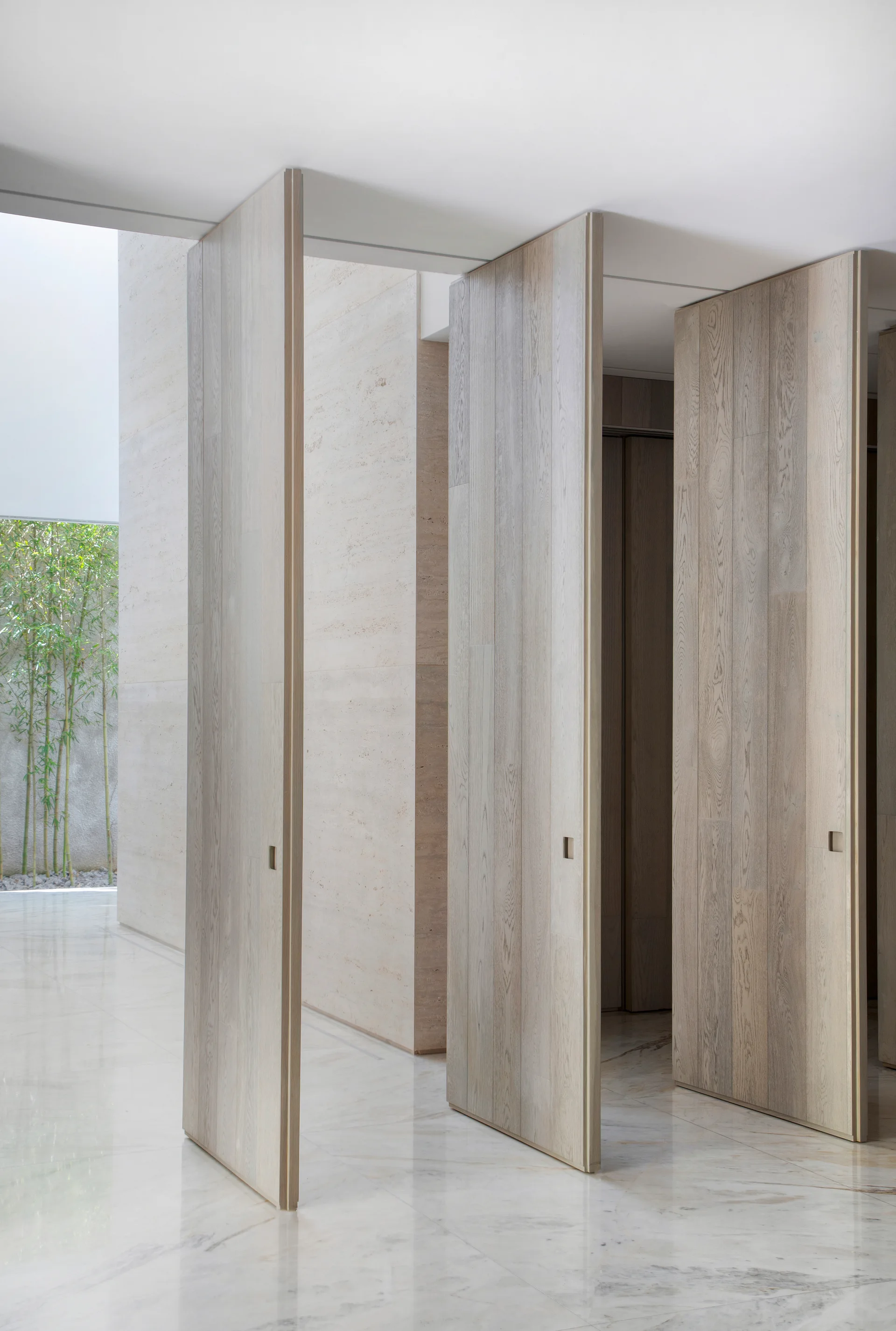 Three tall, light wood pivot doors stand partially open in a minimalist corridor with polished marble flooring and a stone clad wall.