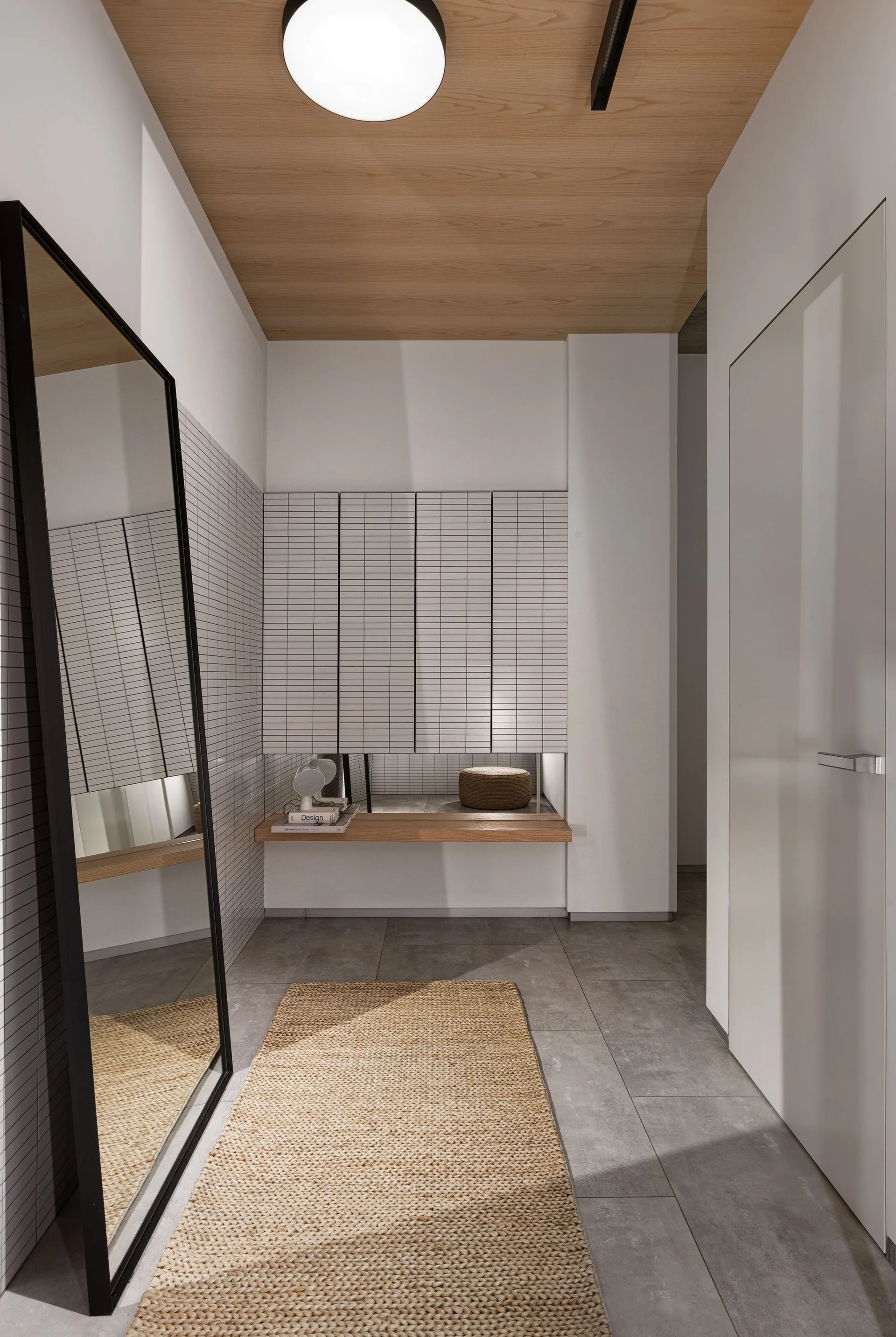 A minimalist entryway with a wood-paneled ceiling, white grid-tiled walls, a tall leaning mirror, and a woven runner rug on gray floor tiles.