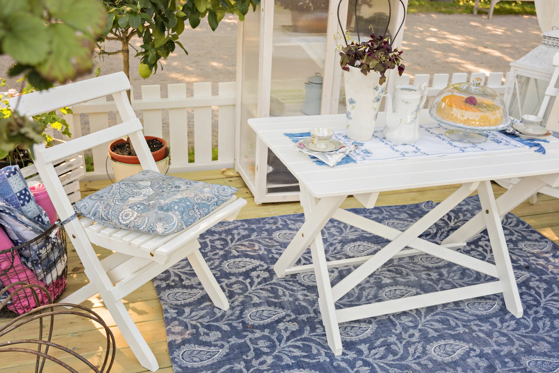 White wooden folding patio furniture stands on a blue patterned rug on a deck next to a white picket fence.