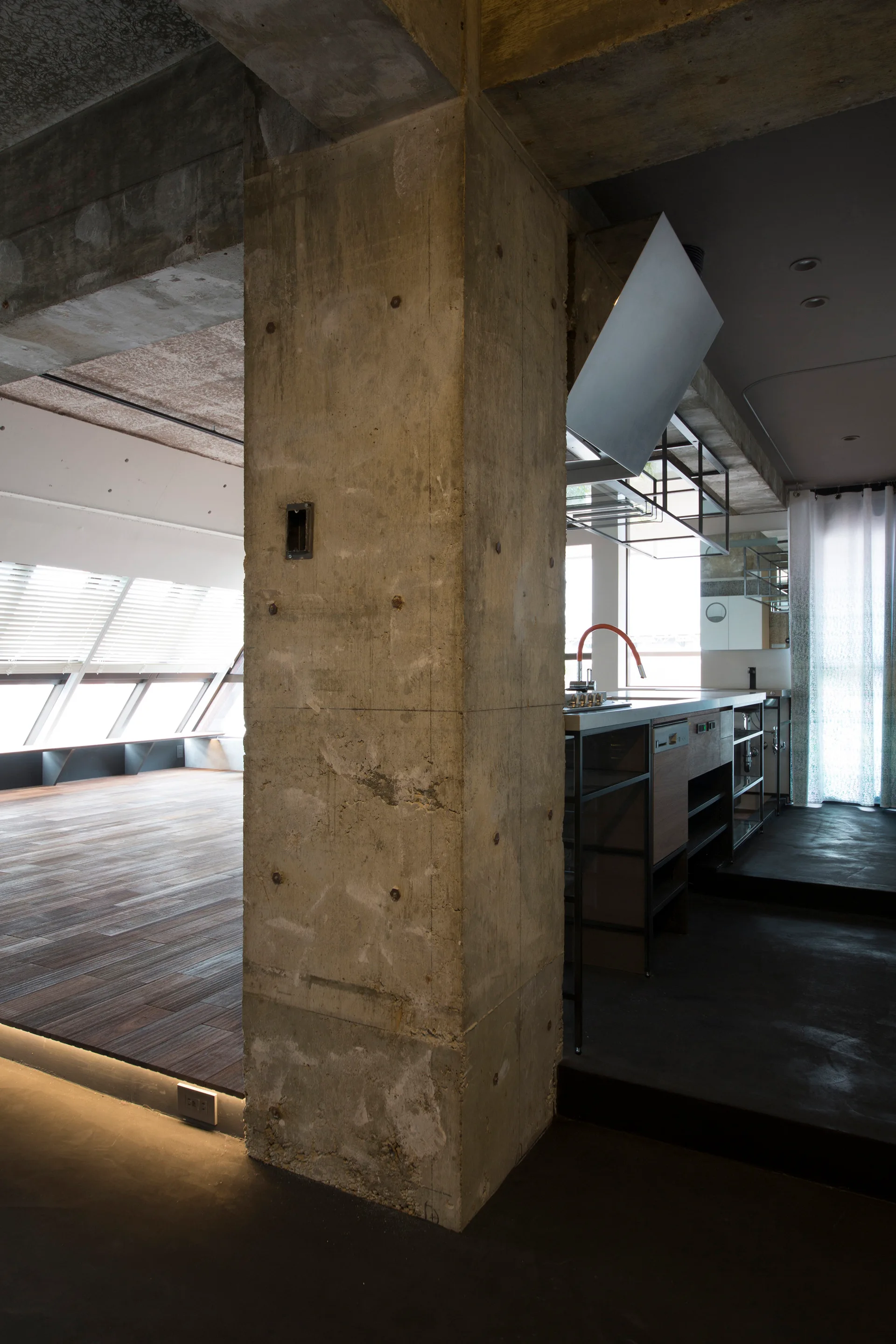 An industrial kitchen featuring a prominent raw concrete column, metal counter, dark wood flooring, and sloped windows in an open floor plan.