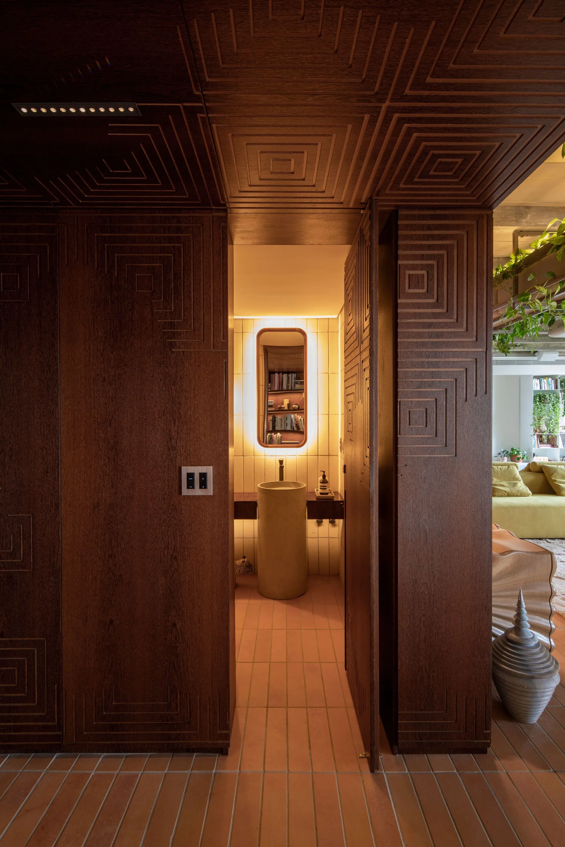 An intricate dark wood paneled doorway with geometric carvings opens to a small powder room featuring a cylindrical stone pedestal sink and a backlit mirror.