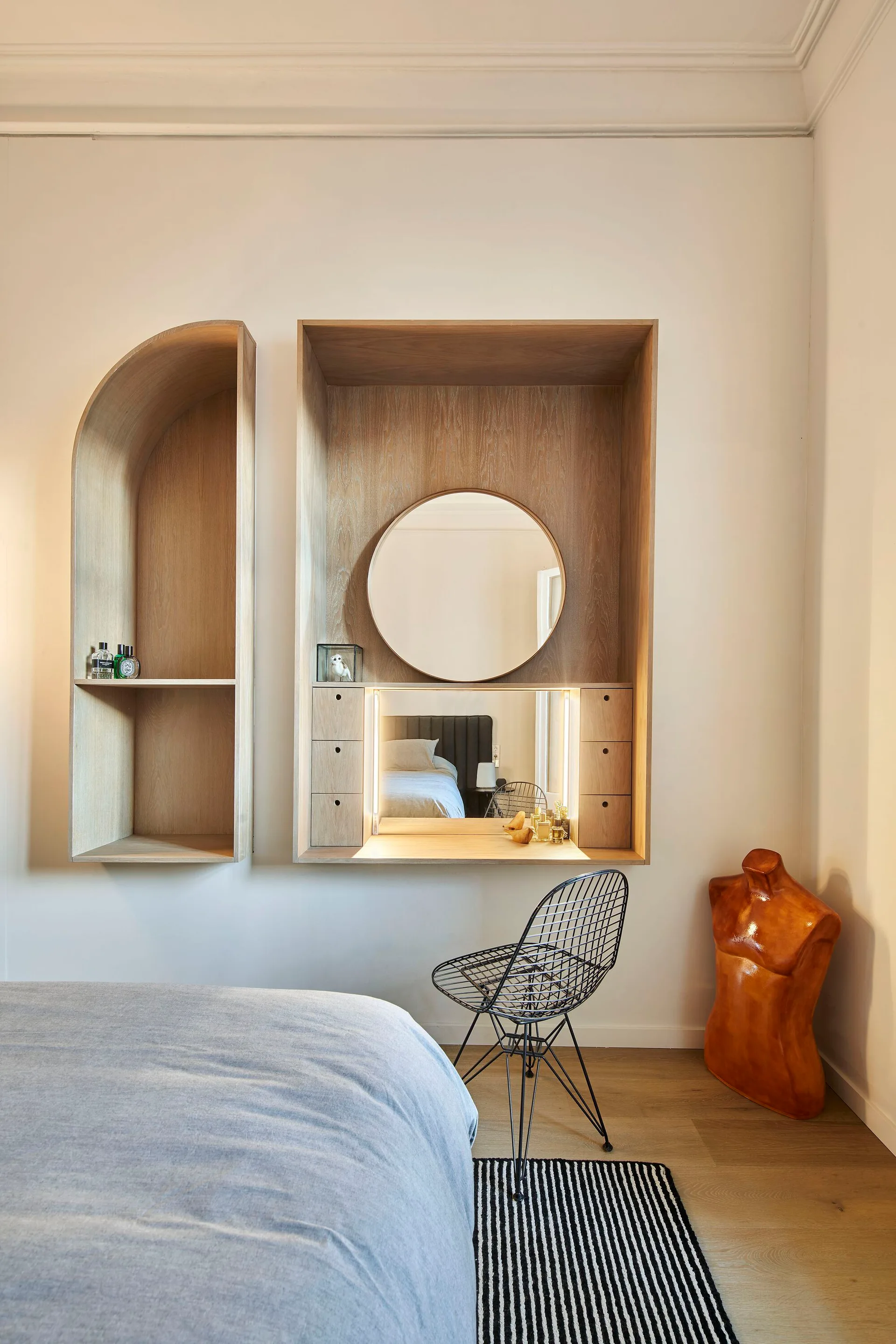 A modern bedroom features a built-in wooden vanity with a round mirror, an arched wall shelf, a black wire chair, and a striped rug.