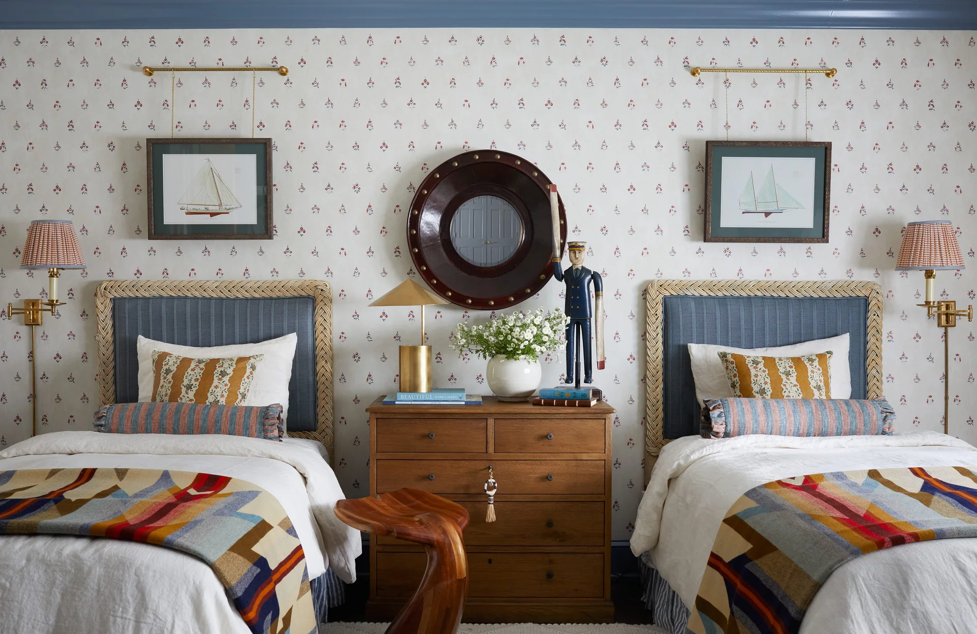 Two twin beds with blue upholstered headboards flank a wooden nightstand under a circular porthole mirror on patterned wallpaper.