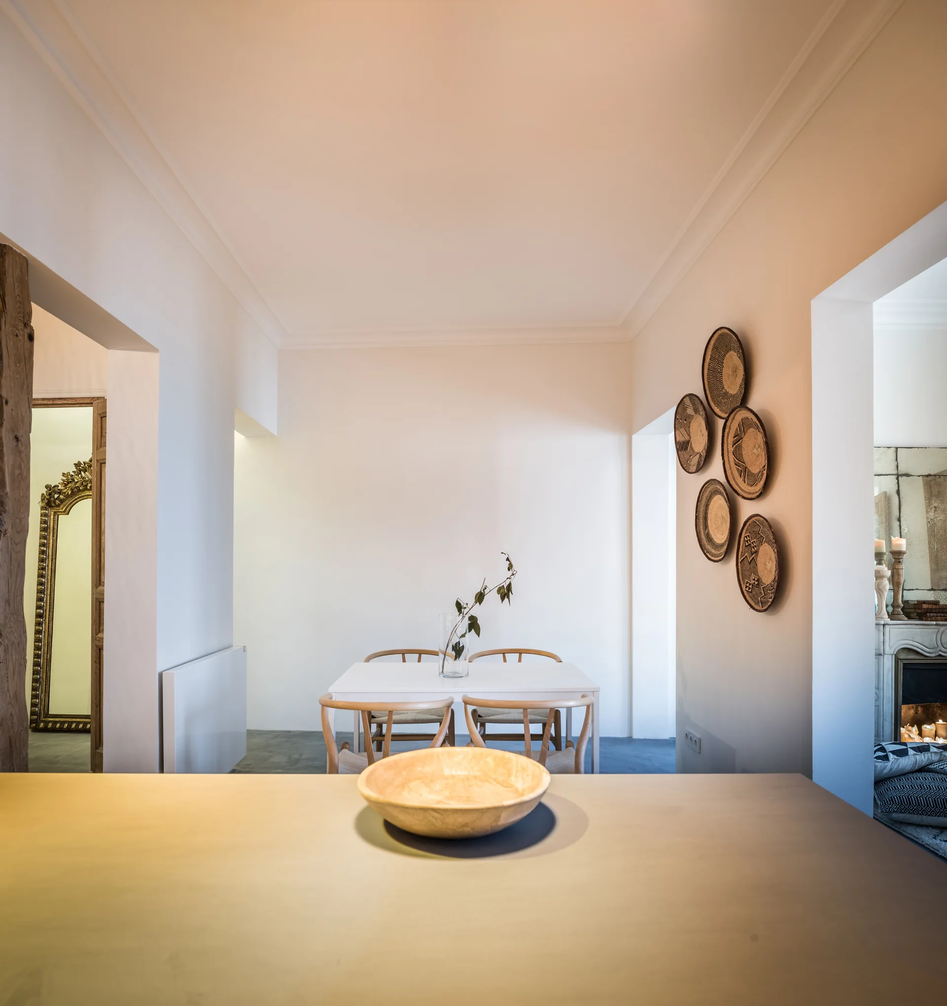A minimalist dining room with a white table, curved wooden chairs, and a cluster of decorative woven baskets on an off-white wall.
