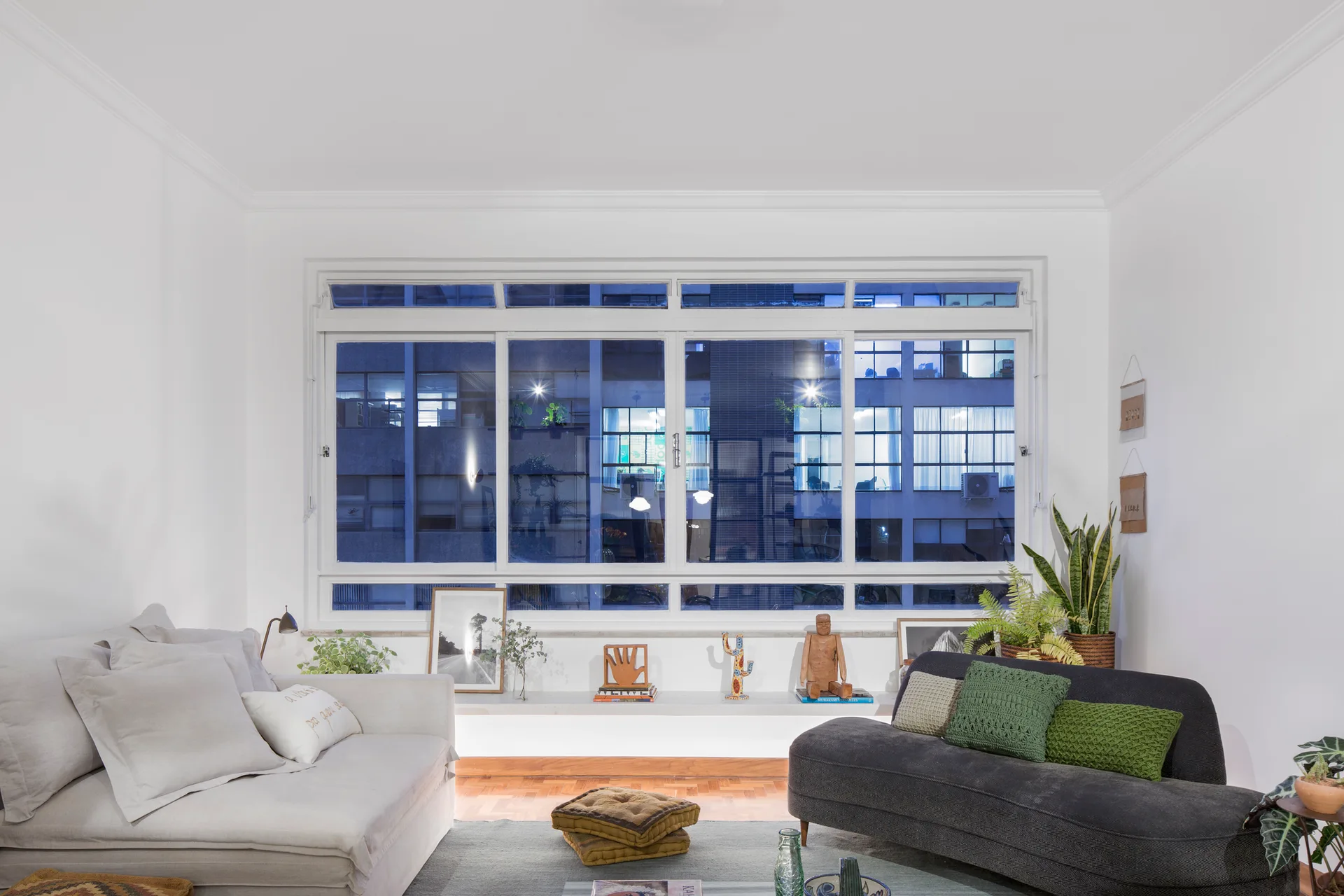 A living room features a light grey sofa and a charcoal grey sofa arranged near a large window with an art-filled ledge.