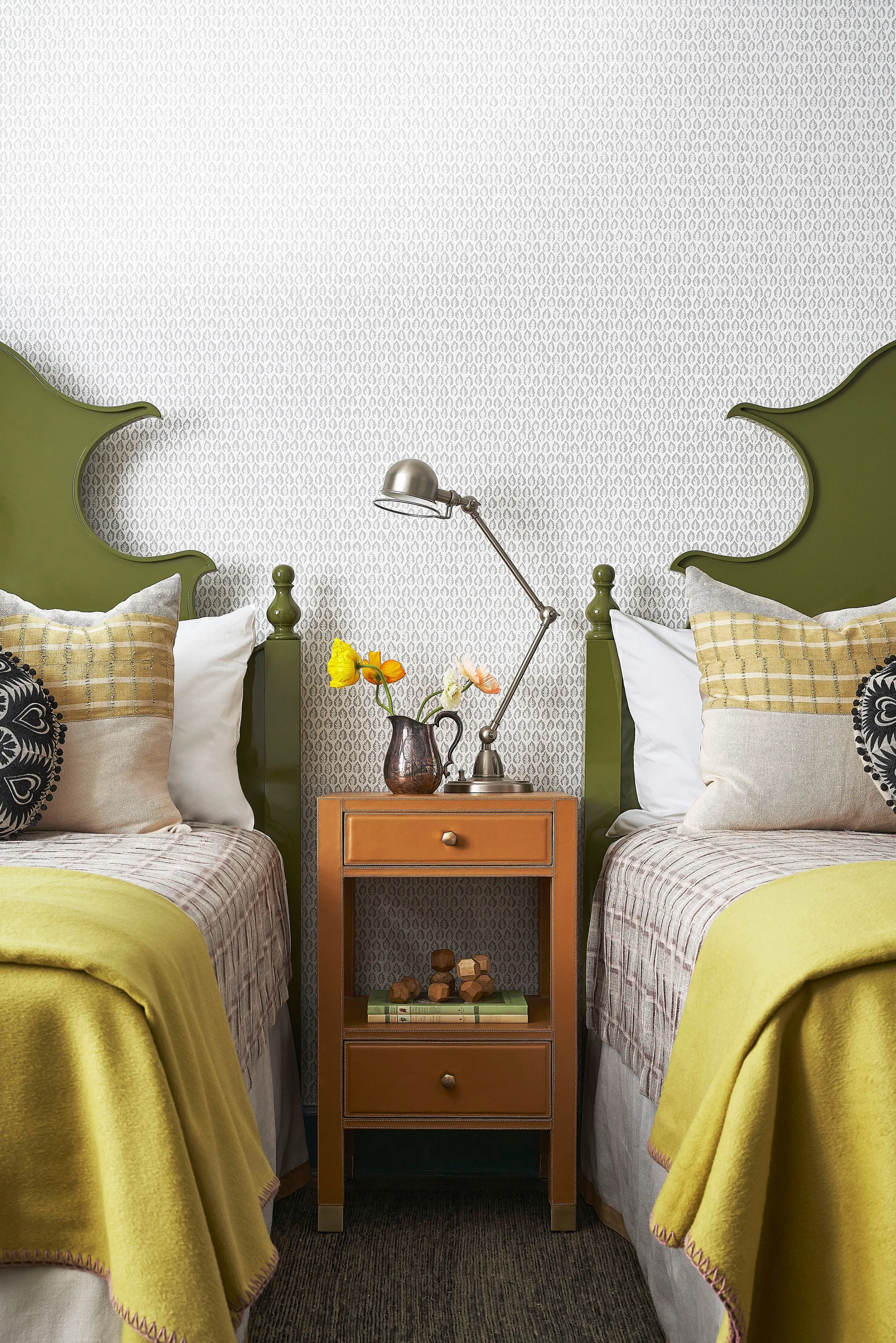 Two twin beds with curved olive green headboards flank a tan leather nightstand in front of light patterned wallpaper.