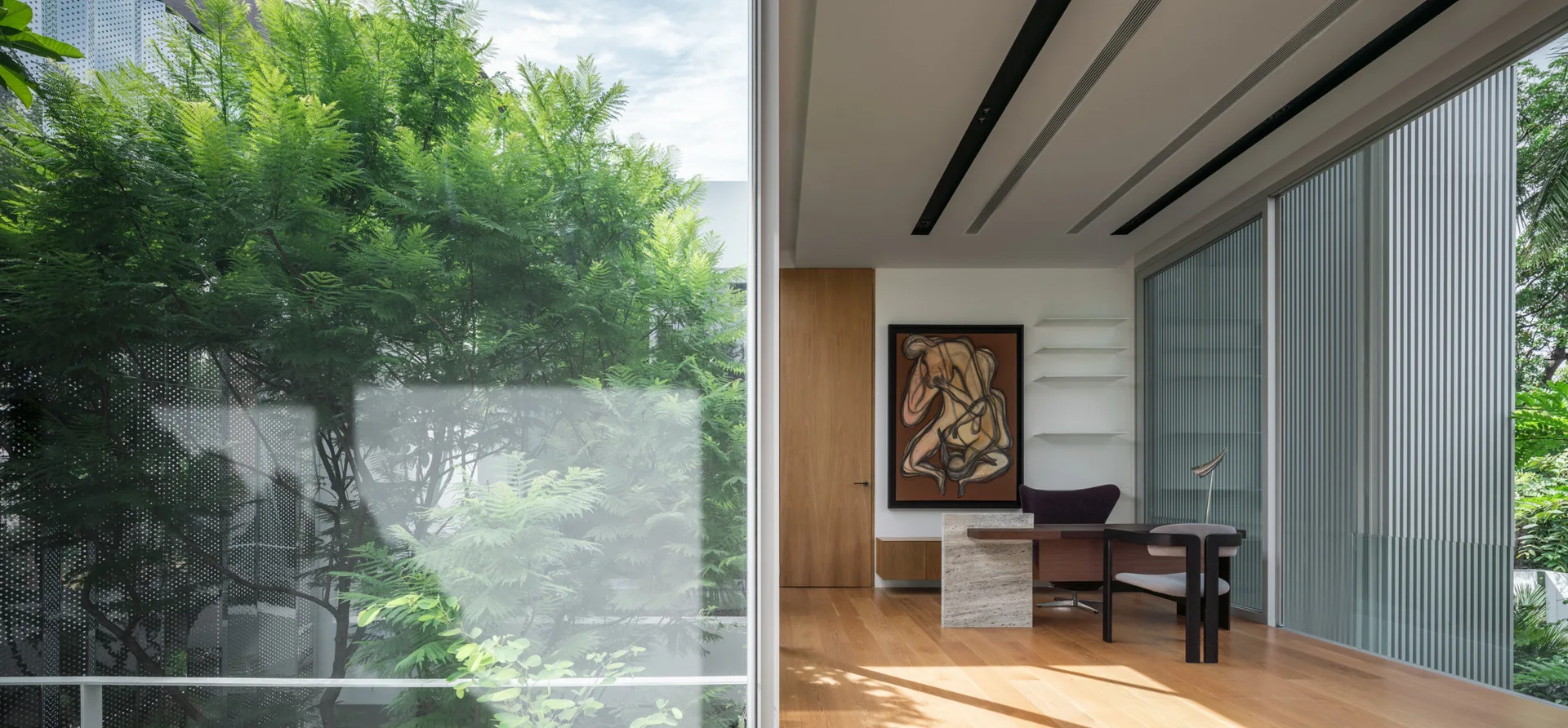 A minimalist home office featuring a desk with a stone pedestal base, wood flooring, and large glass windows overlooking dense green foliage.