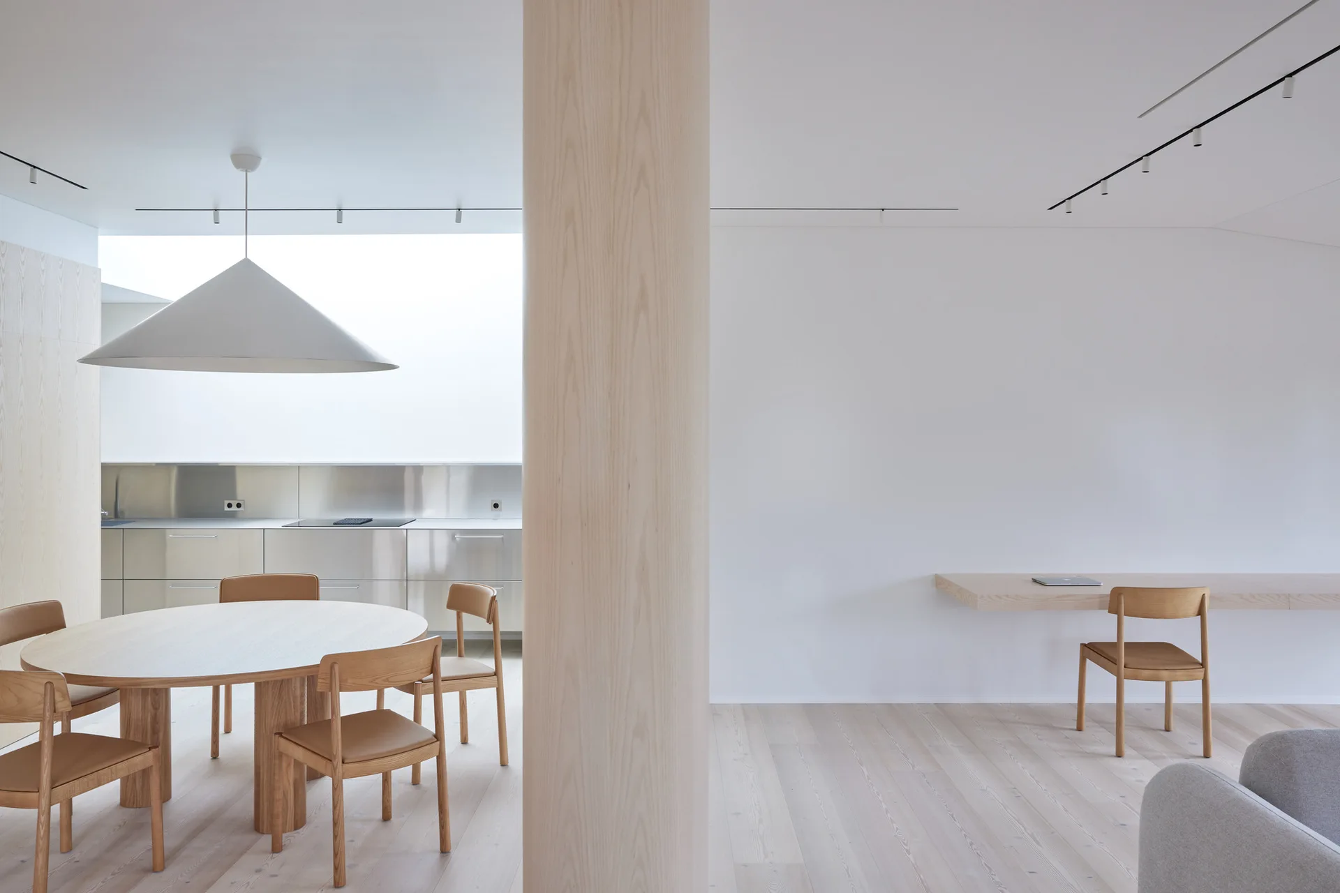 A minimalist dining room featuring a large light wood column, a circular wooden table with chairs, and a stainless steel kitchen in the background.