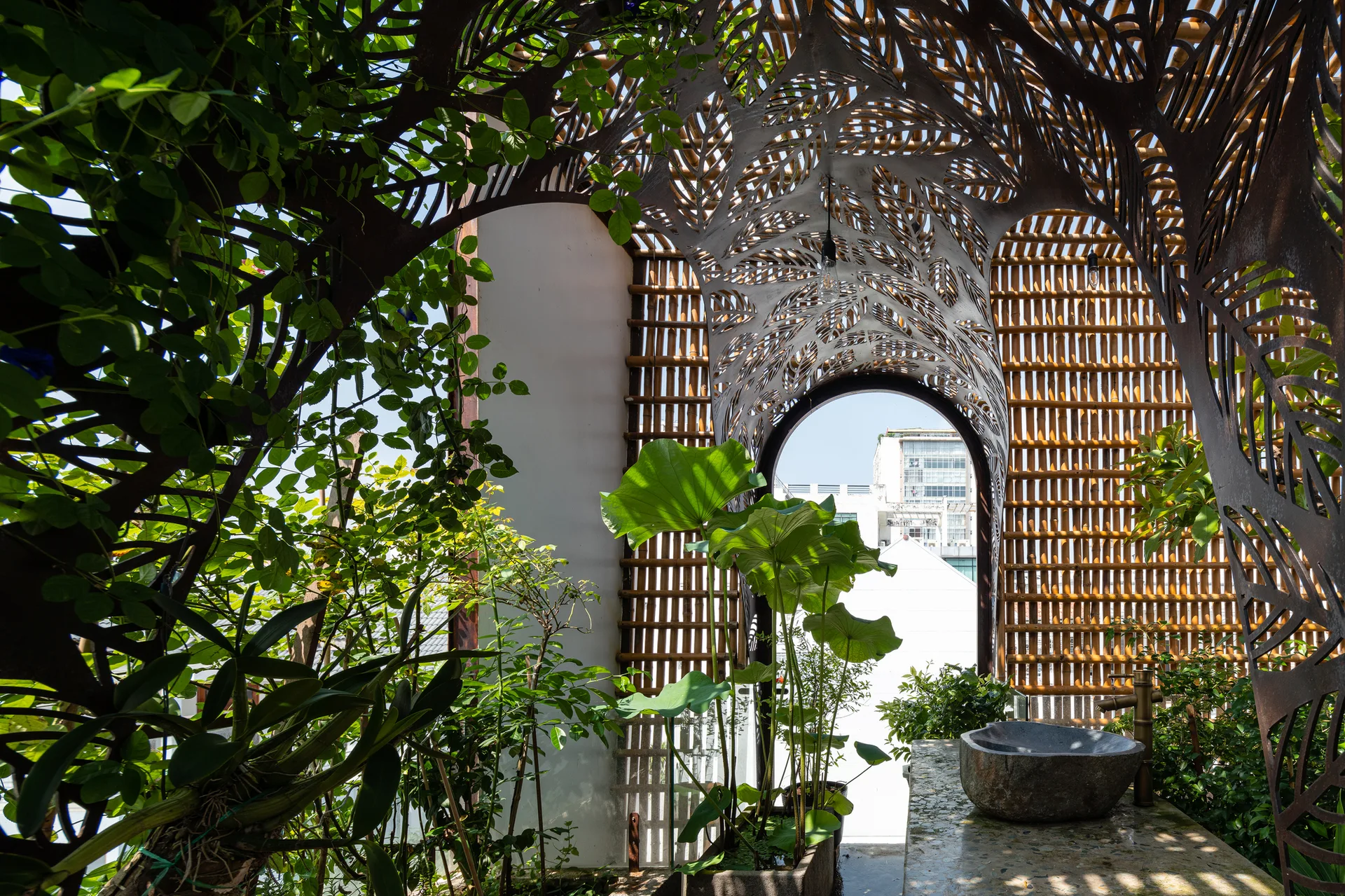 An outdoor garden features an ornate laser-cut metal canopy over a stone basin with vertical bamboo screens and lush potted plants.