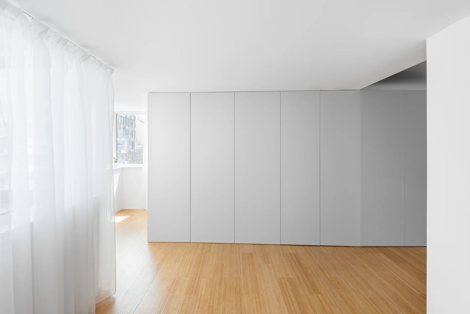 A minimalist room with a seamless white built-in wardrobe wall, light wood plank flooring, and a sheer white curtain filtering natural light.
