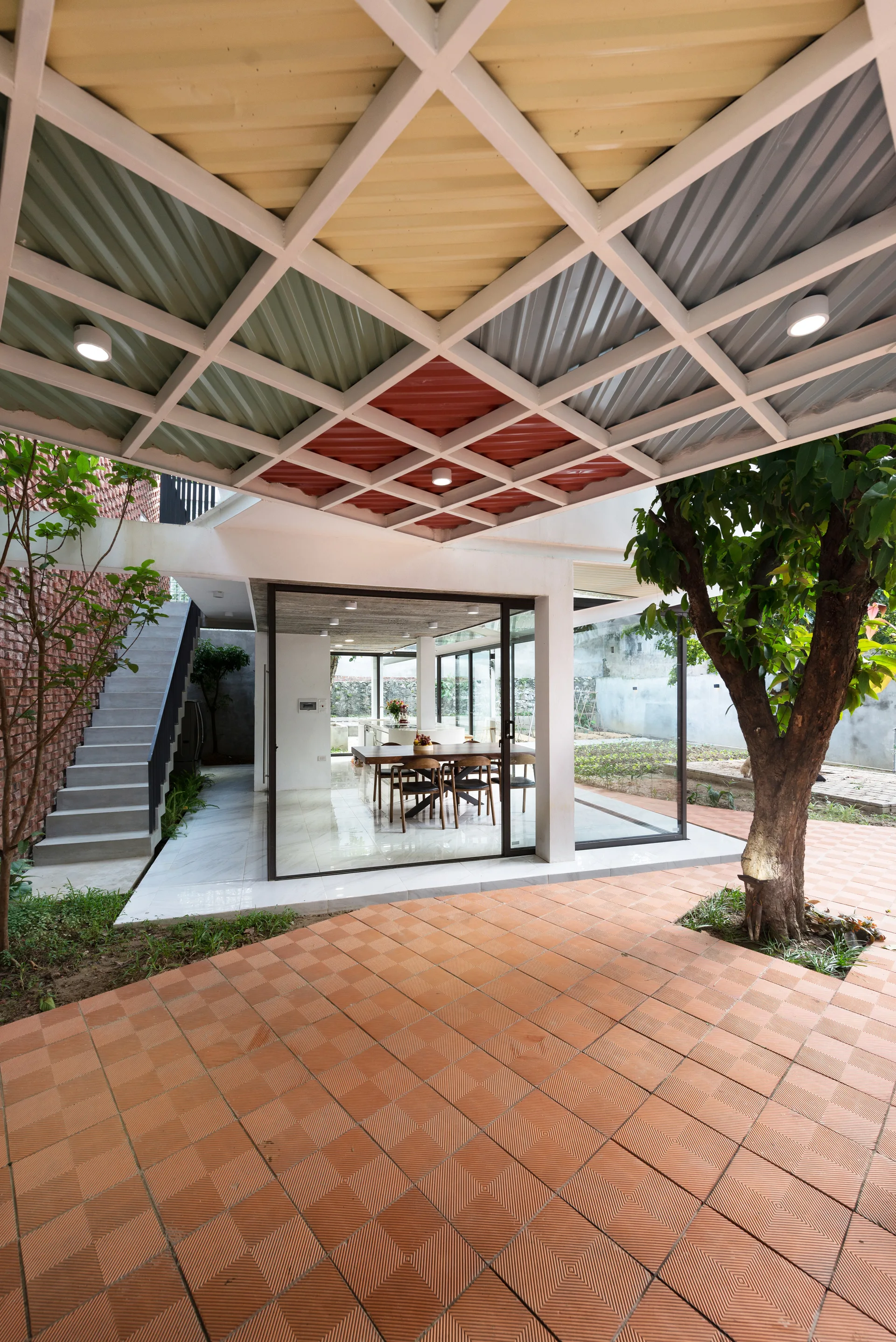 A modern outdoor patio with textured terracotta tiles and a multicolored geometric grid canopy leading to a glass-enclosed dining space.