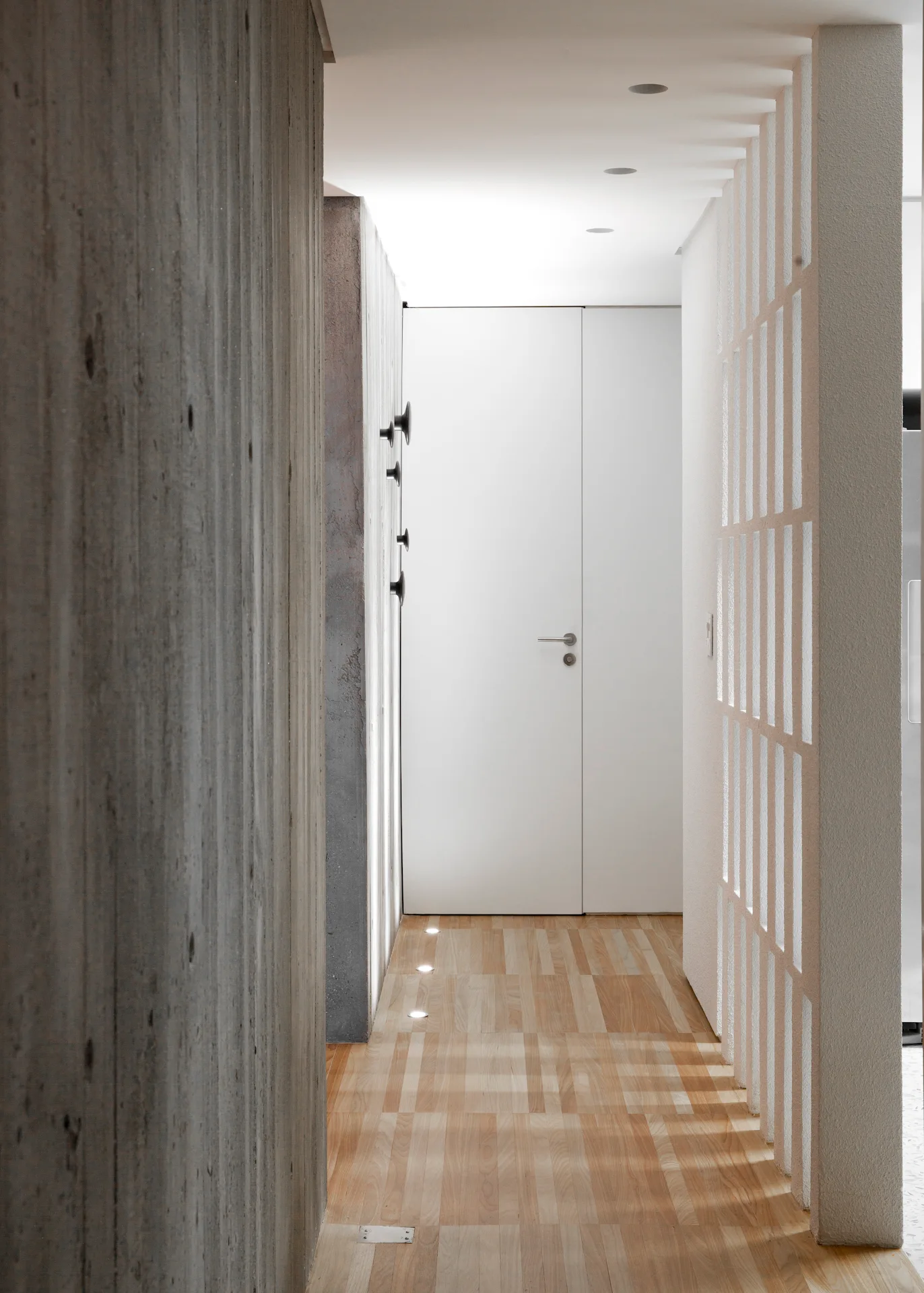 Modern corridor featuring a textured board-formed concrete wall, a white slatted partition, parquet flooring, and a white door at the end.