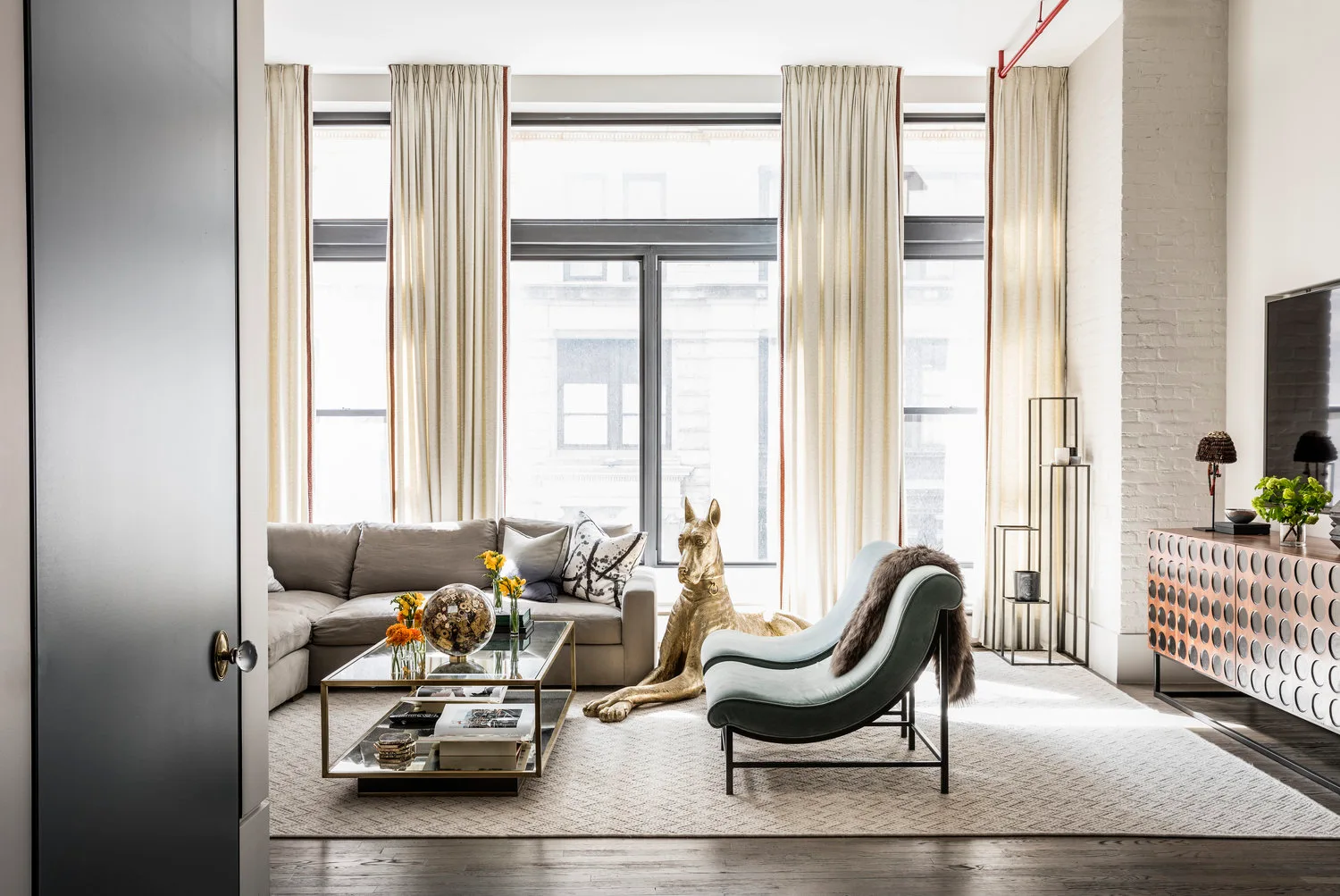 A bright loft living room features a grey sectional, a curved mint green accent chair, a gold dog statue, and a textured wooden sideboard.