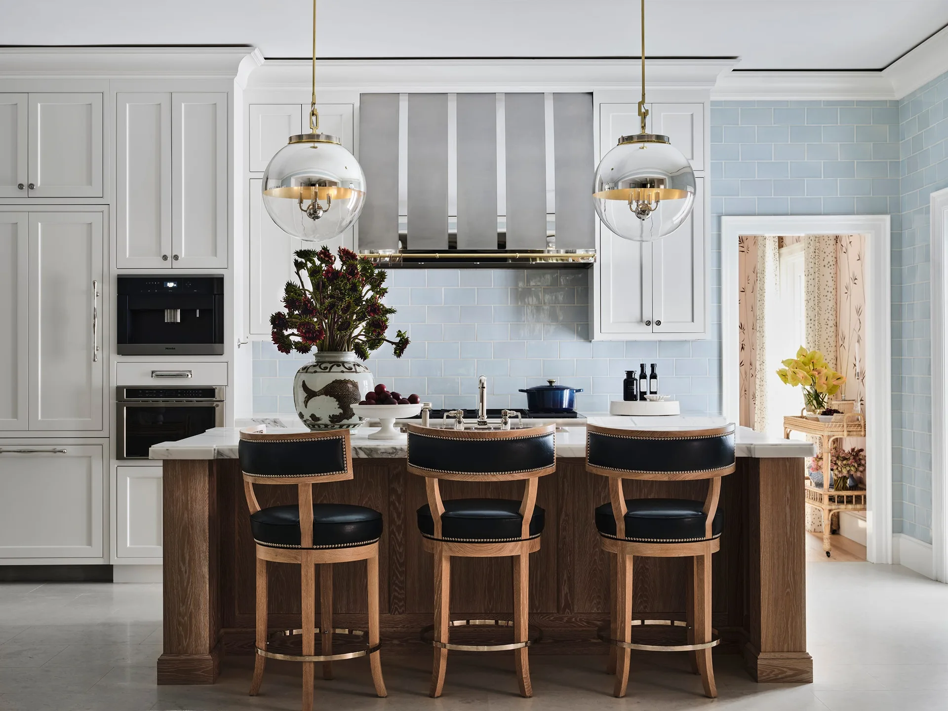 A kitchen featuring a wood island with three black leather stools, a light blue tiled backsplash, and white cabinetry under sphere glass pendants.
