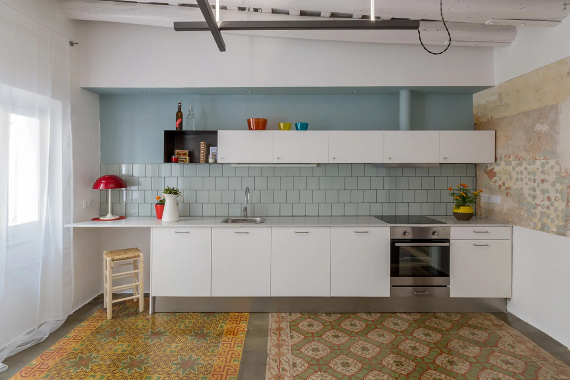 A linear kitchen featuring white cabinetry, a pastel blue tiled backsplash, white ceiling beams, and multi-patterned colorful floor tiles.