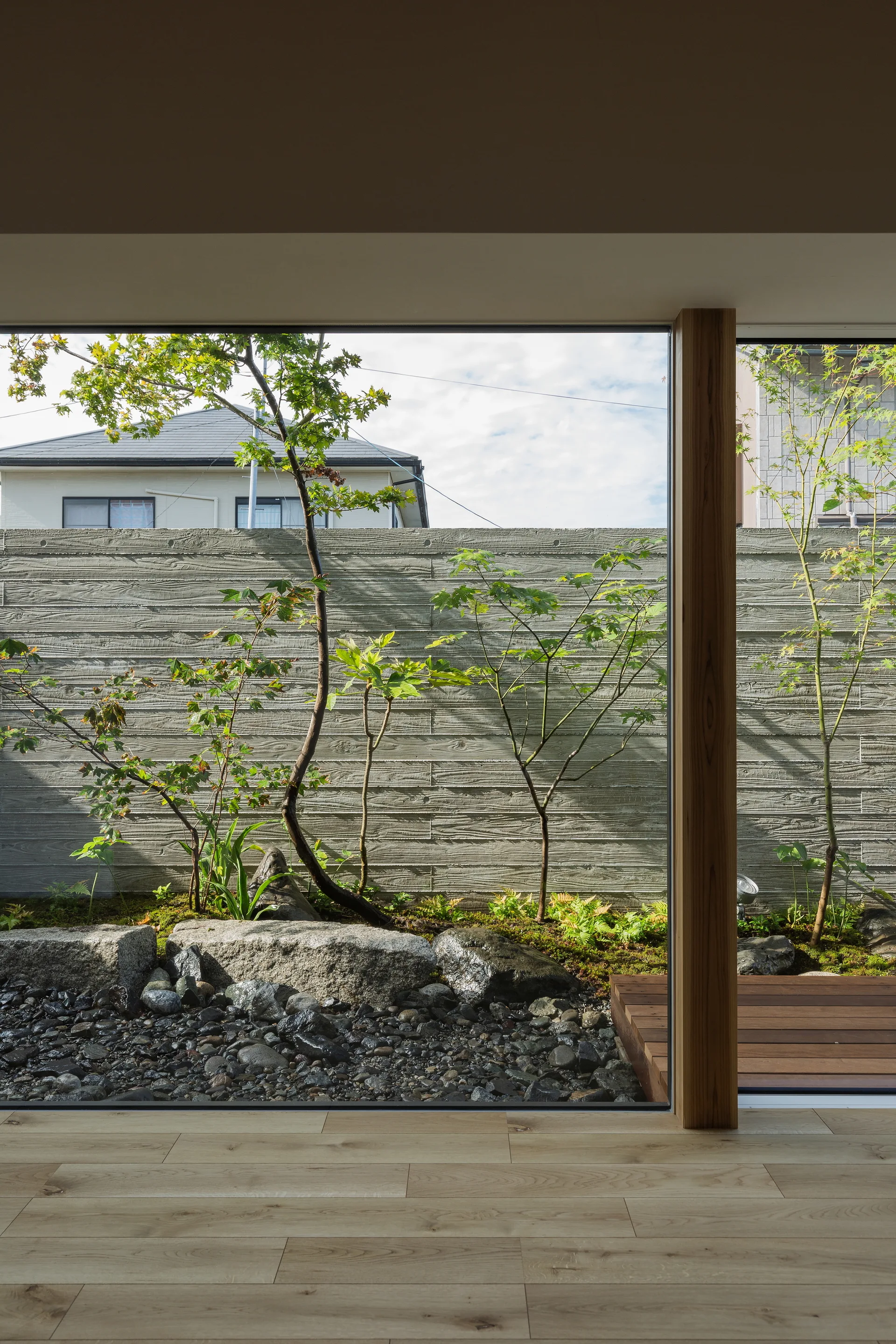 A large picture window looks out onto a landscaped garden featuring a board-formed concrete retaining wall, stone boulders, and small trees.