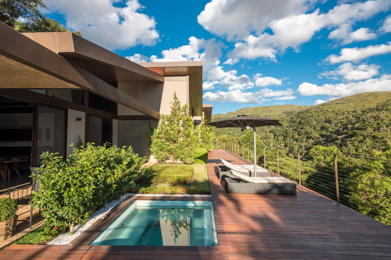 A spacious wooden deck features a small square plunge pool, two loungers with an umbrella, and a metal cable railing overlooking a forested mountain valley.