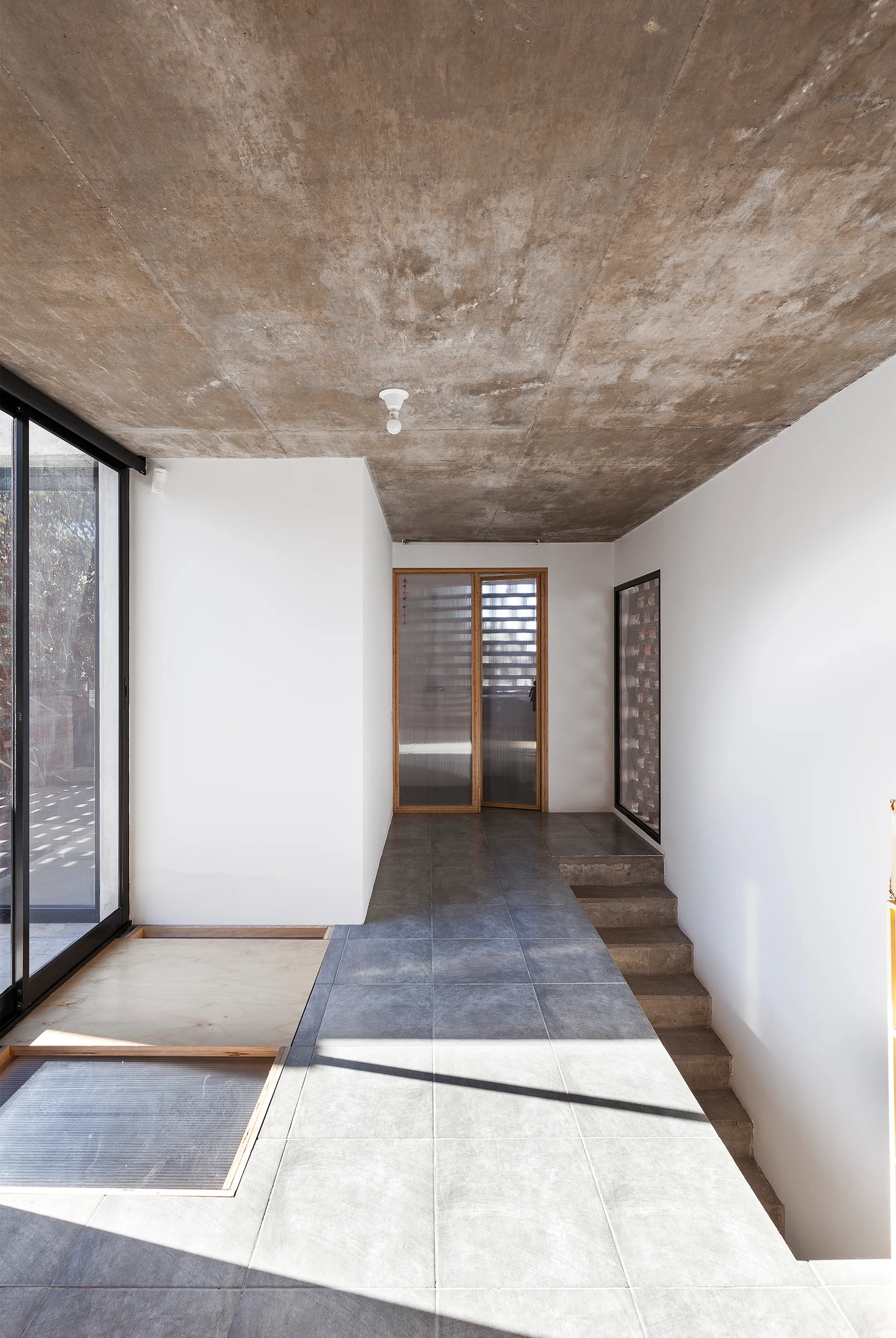 A minimalist entryway featuring an exposed concrete ceiling, grey tile flooring, and a wood-framed fluted glass door leading to concrete steps.