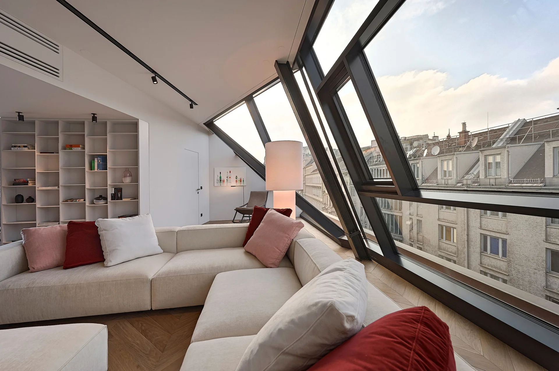 A modern living room features a cream sectional sofa on herringbone wood floors next to a large slanted architectural window wall.