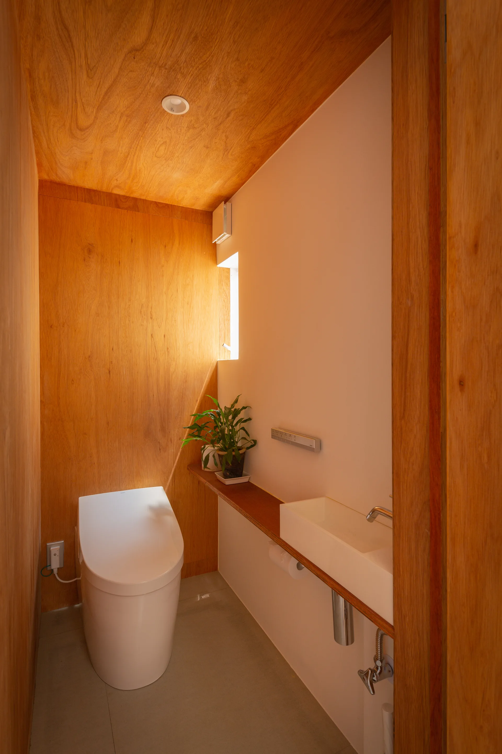 A narrow minimalist powder room features warm wood-paneled walls and ceiling, a white toilet, and a slim floating sink over a concrete floor.
