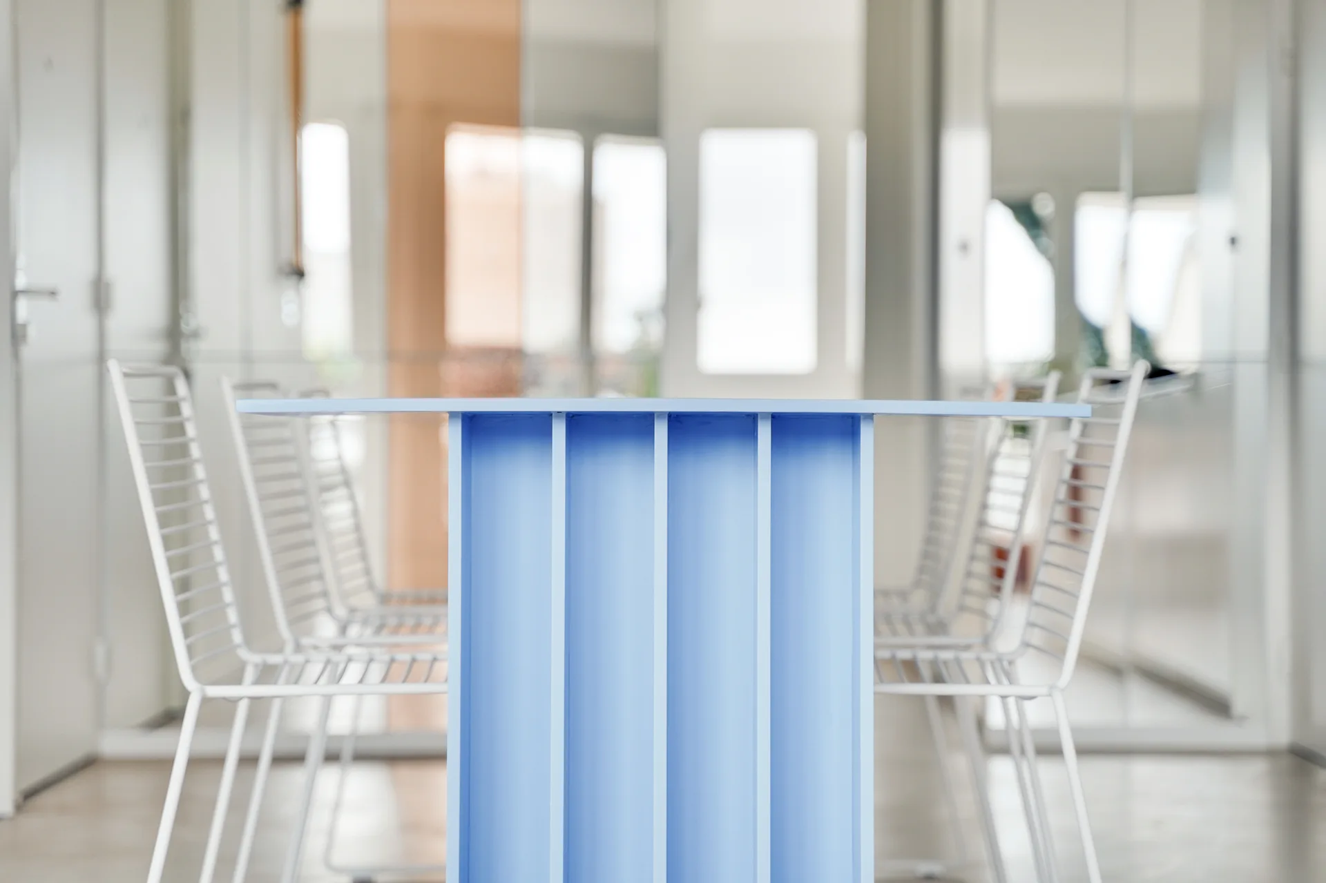 A light blue dining table with a ribbed sculptural base is surrounded by white wire chairs in a bright interior space.