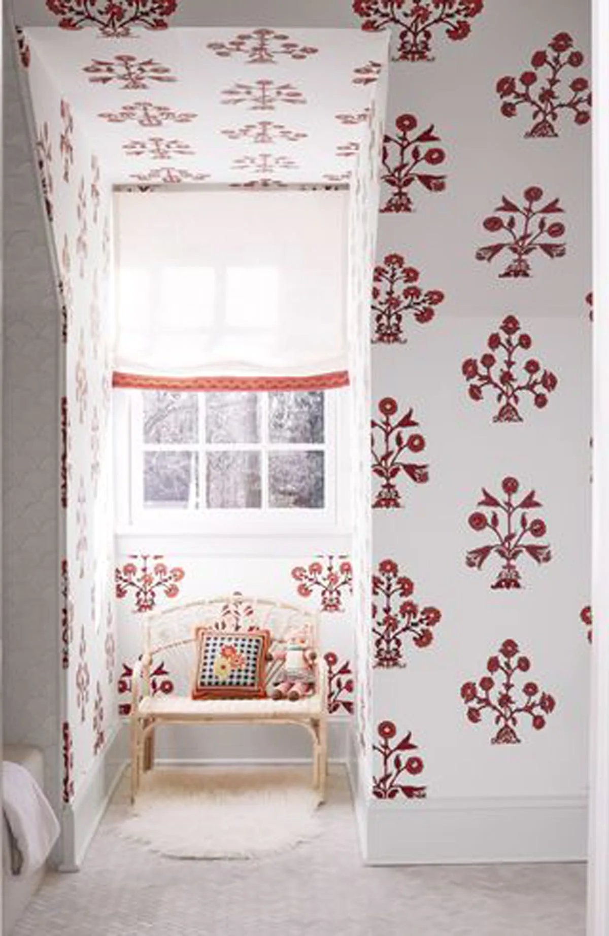 A small window alcove covered in red and white floral wallpaper featuring a light wood wicker bench with a patterned cushion.
