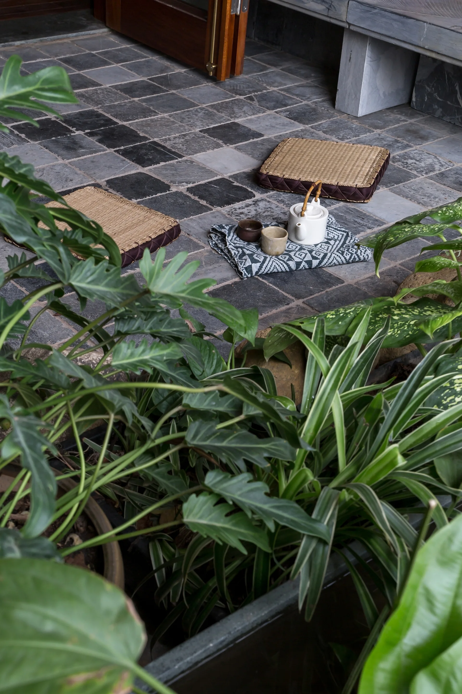 Two square straw floor cushions and a white ceramic tea set sit on a grey stone tiled patio surrounded by lush green plants.