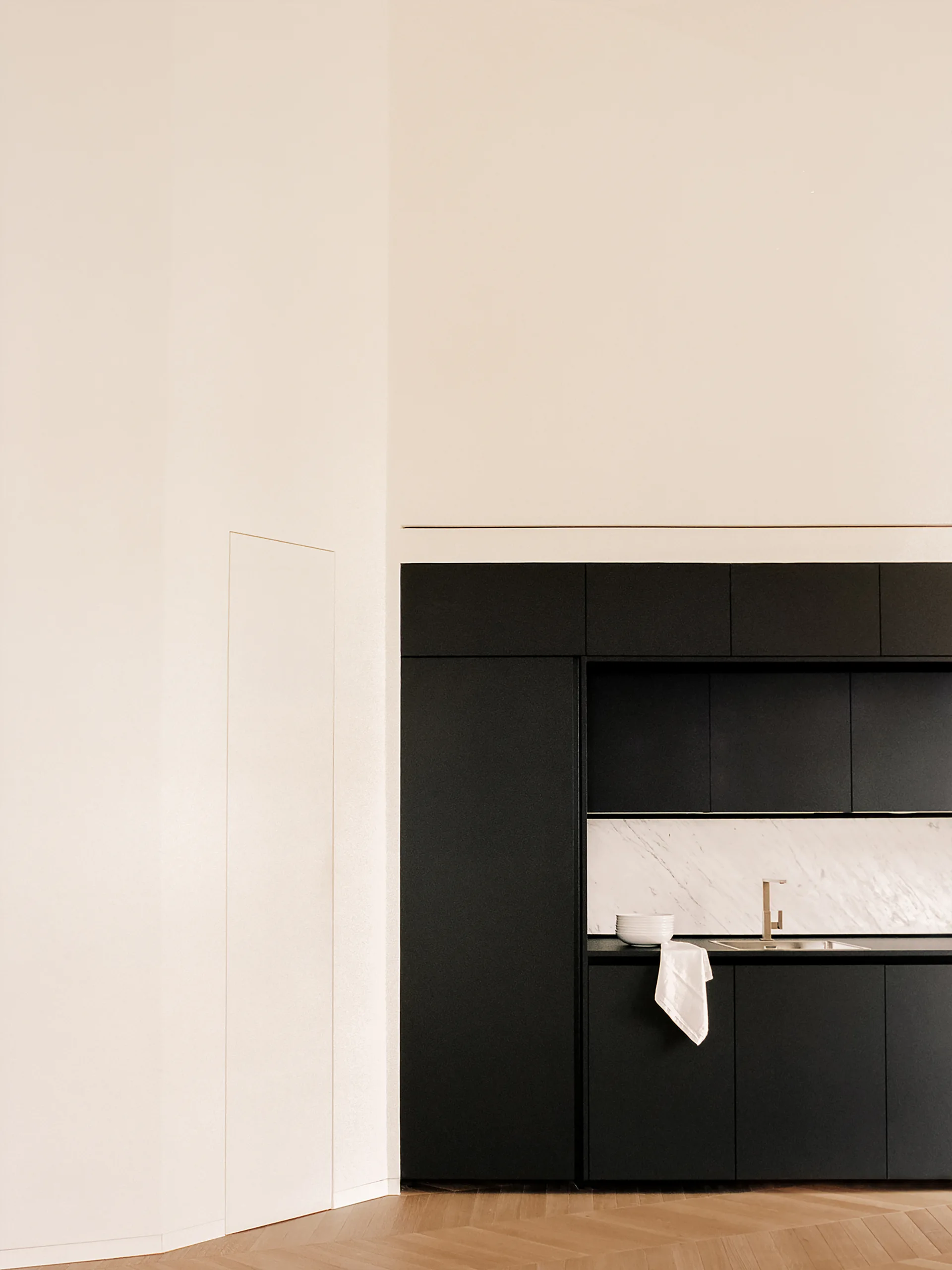 Matte black kitchen cabinetry built into a beige wall features a white marble backsplash, a gold faucet, and herringbone wood flooring.