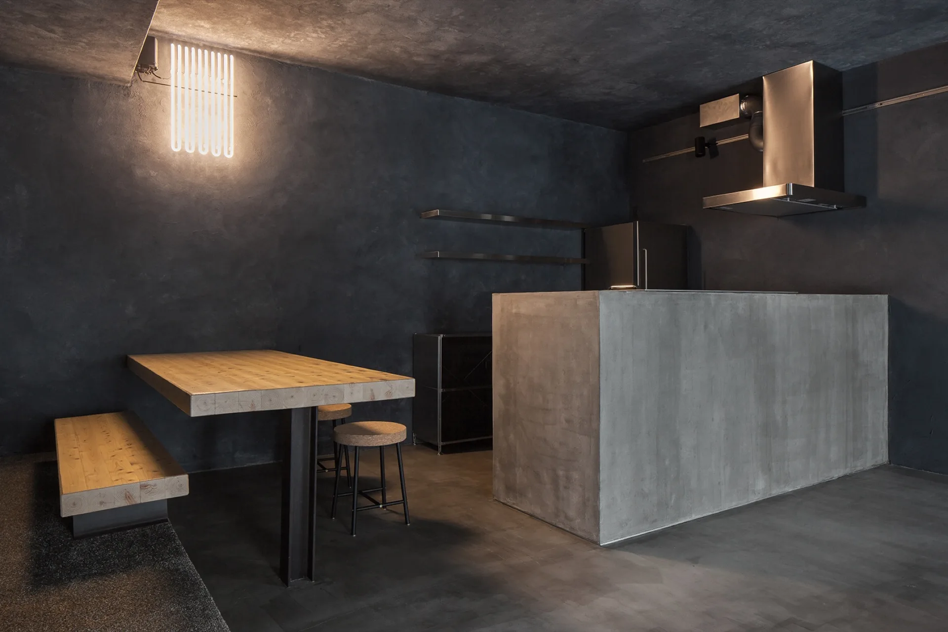 A dark minimalist kitchen features a monolithic concrete island, textured charcoal walls, and a thick light wood dining table with matching bench.