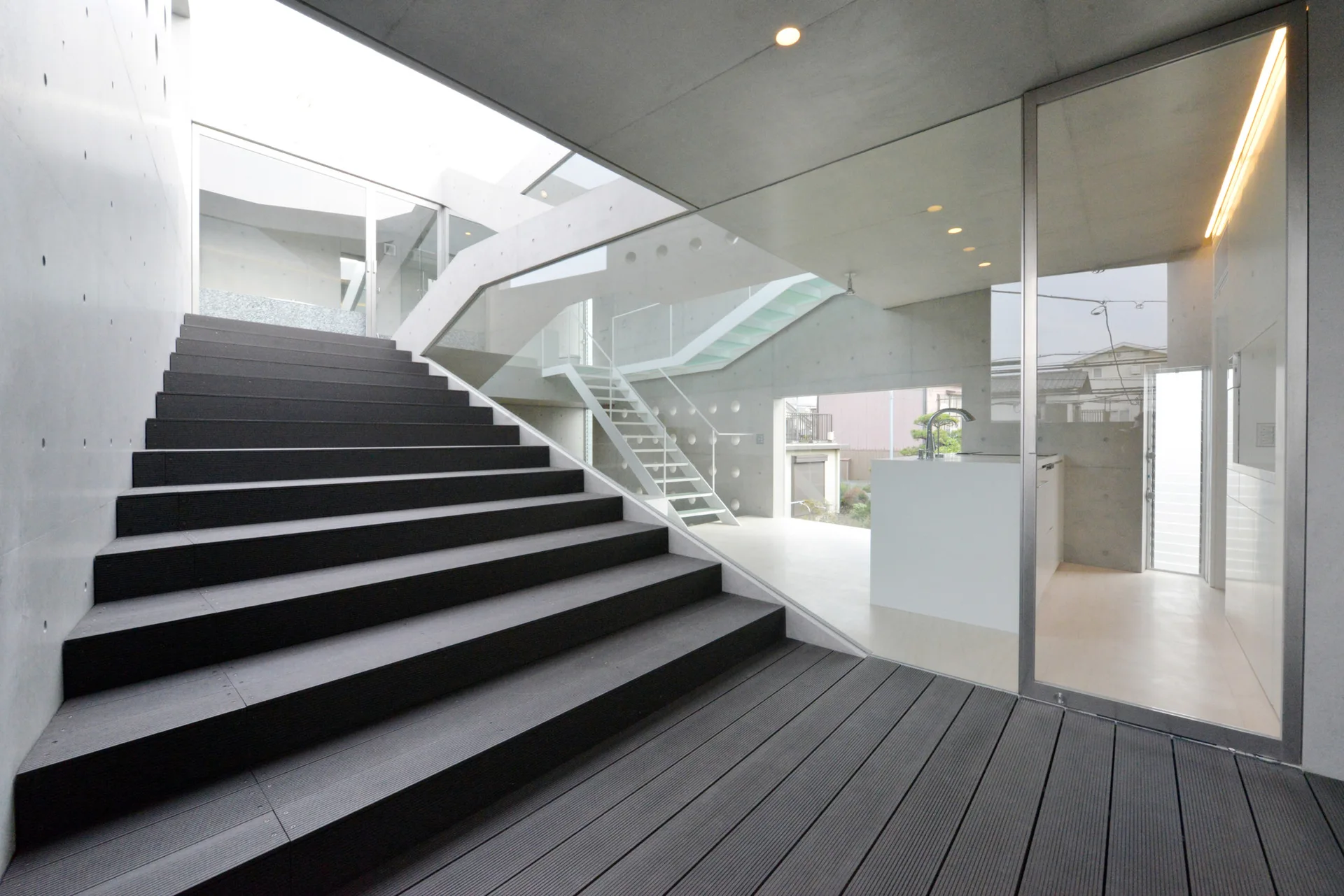 A wide dark wood staircase leads up past concrete walls and a large glass partition that reveals a white kitchen island.