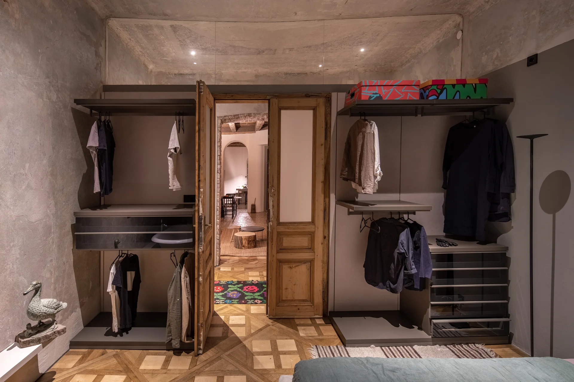 An open modular closet system with hanging clothes and drawers stands on a geometric parquet wood floor flanking a rustic wooden doorway.