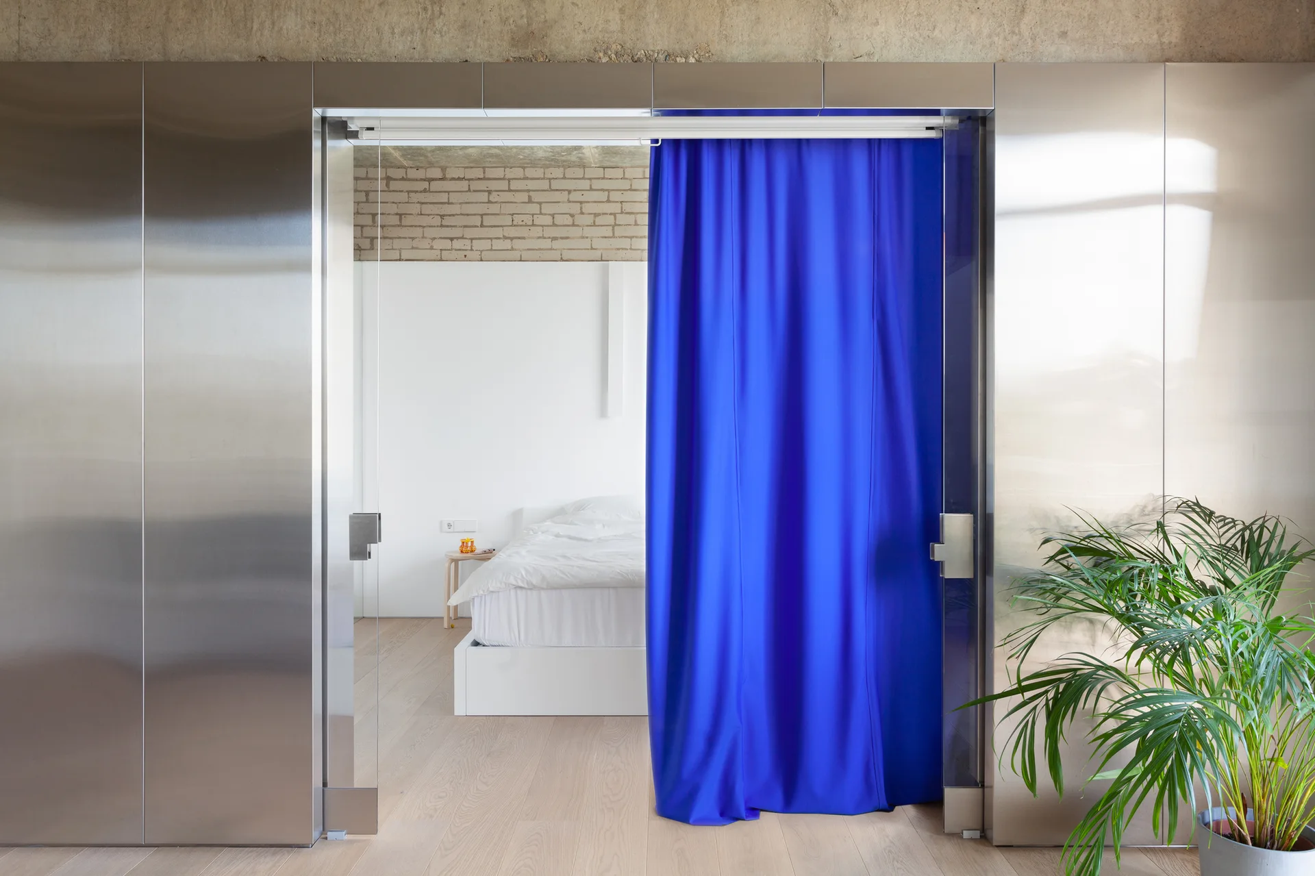 A stainless steel doorway features a vibrant blue curtain partially drawn, showing a minimalist bedroom with a white bed and exposed brick.